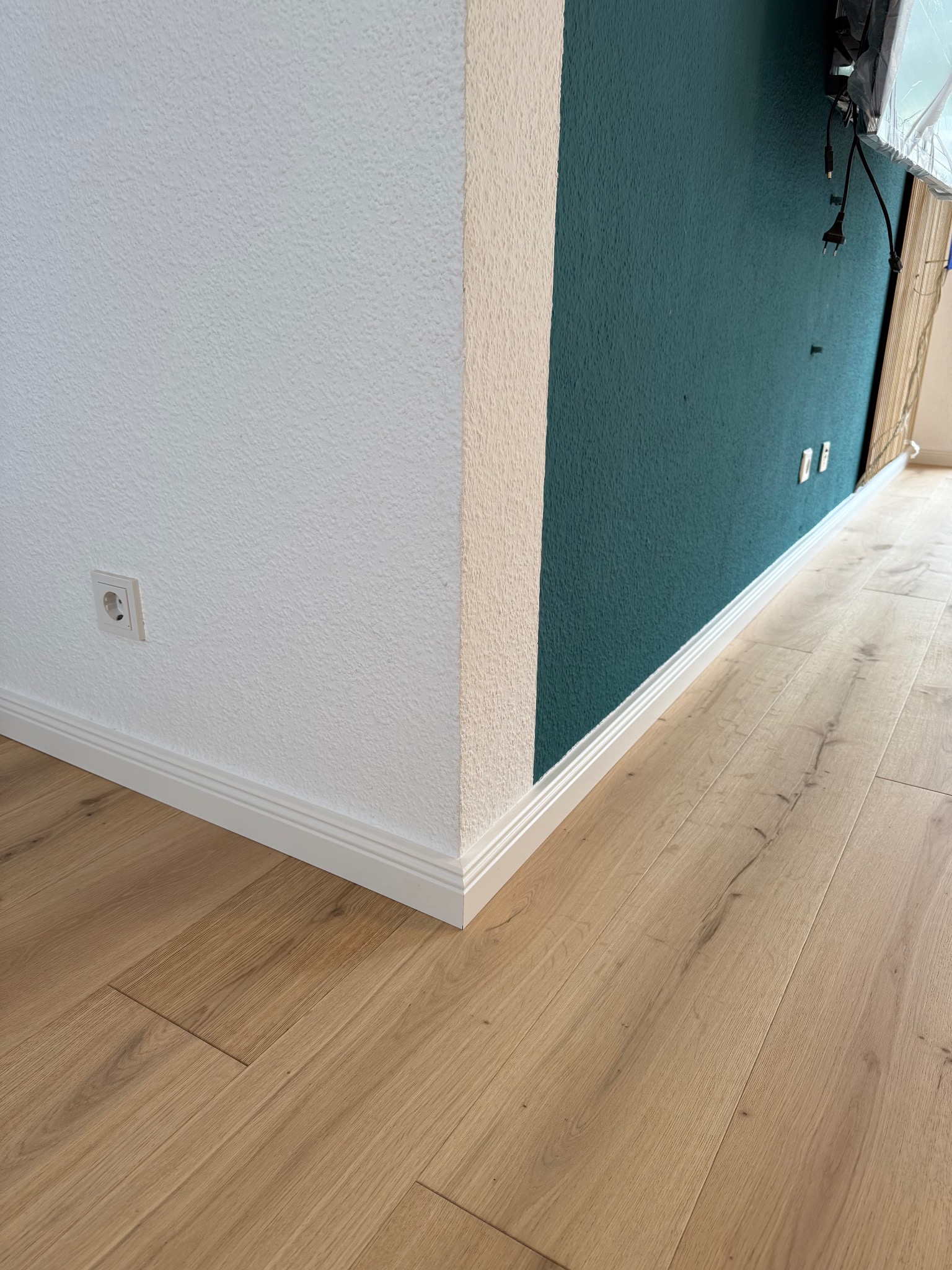 Jasne panele podłogowe z listwami przypodłogowymi, stykające się z białą i zieloną ścianą o wyraźnej fakturze, gniazdko elektryczne przy podłodze.