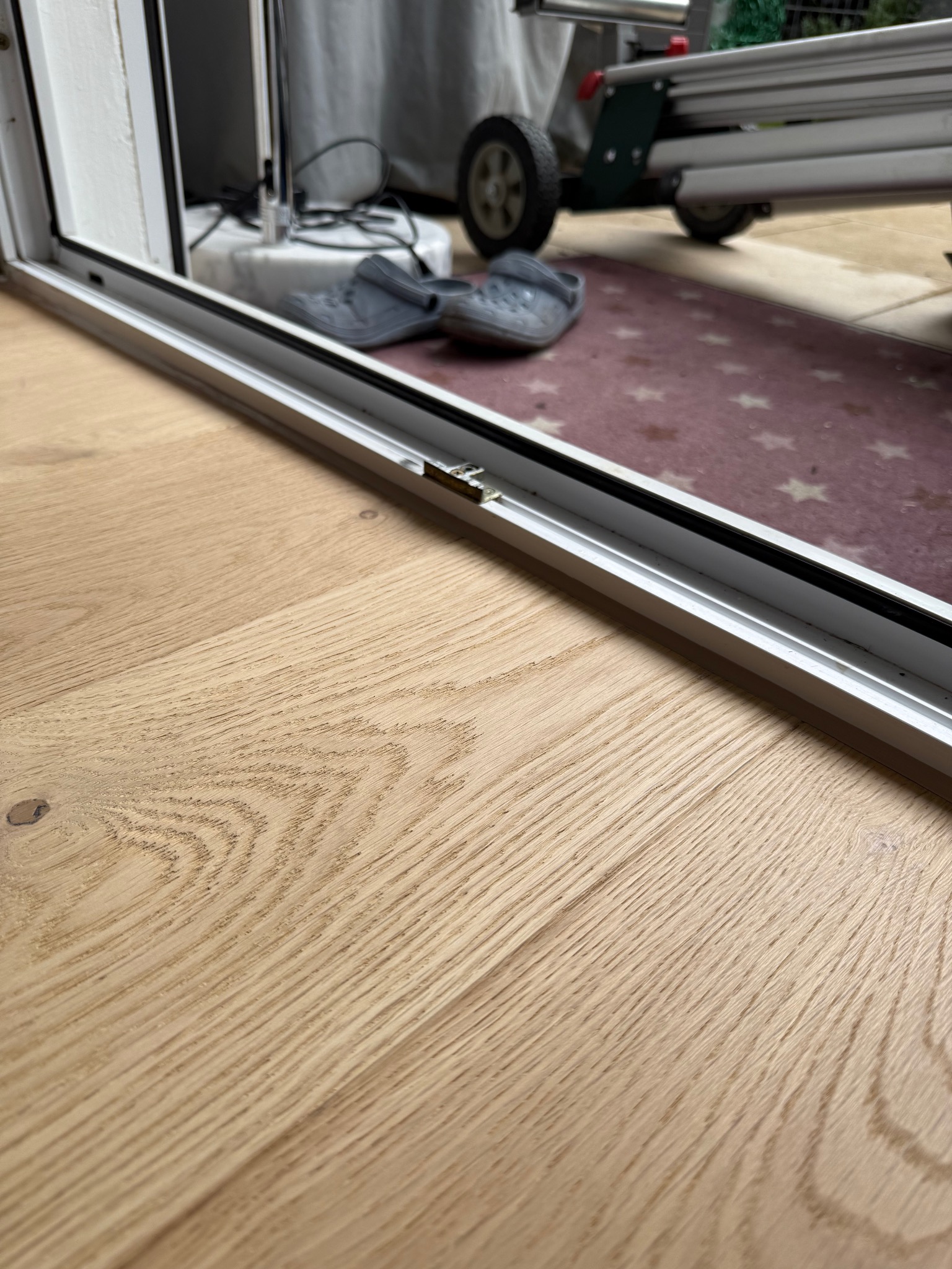 Jasny, drewniany parkiet ułożony przy białej ramie okiennej. W tle widoczne szare klapki i fragment maszyny do obróbki podłóg na dywaniku w gwiazdki.