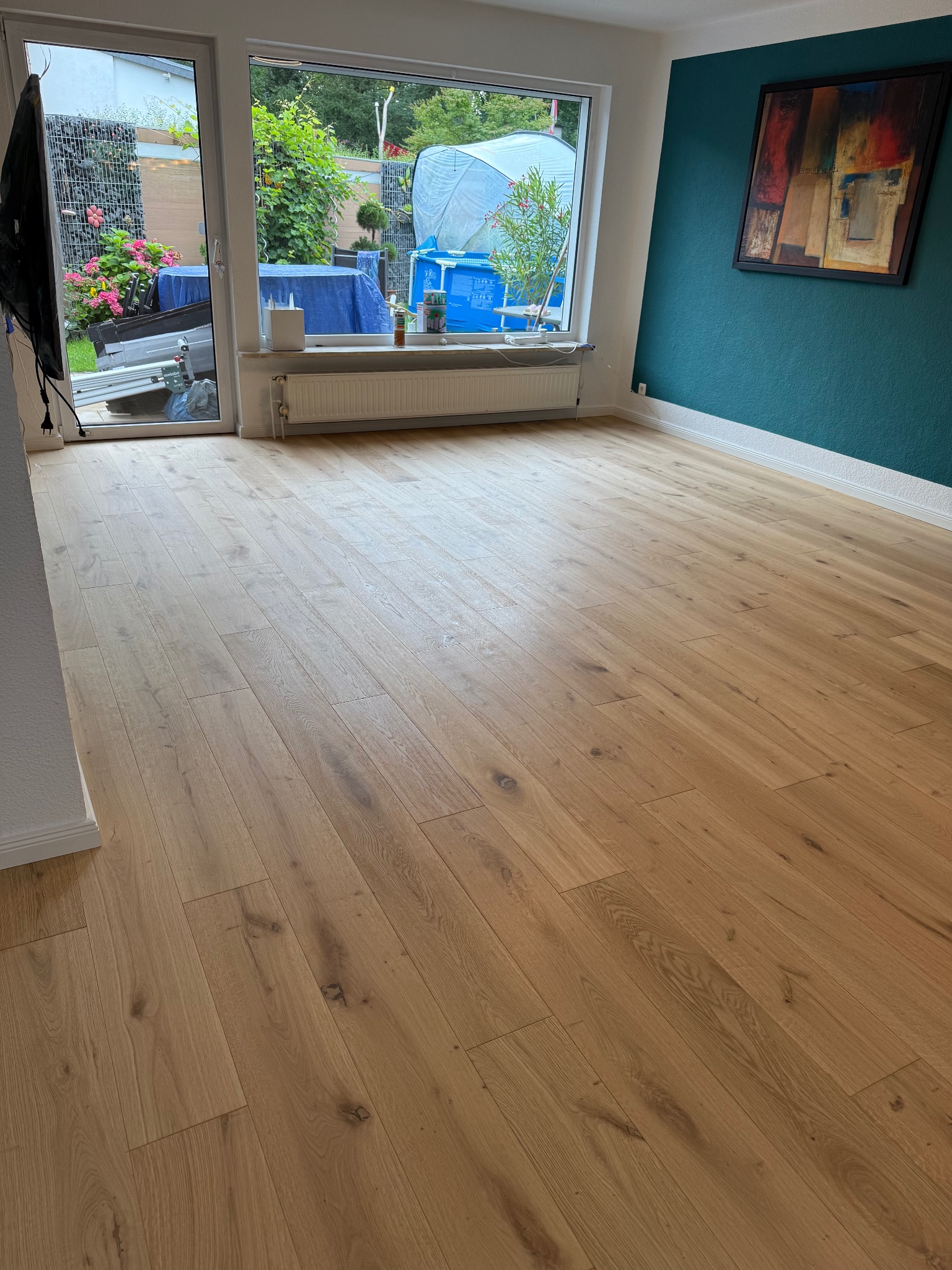 Jasny, wykończony parkiet w nowym wnętrzu z oknem na ogród i turkusową ścianą z obrazem. Widok na ułożone deski podłogowe w pokoju.