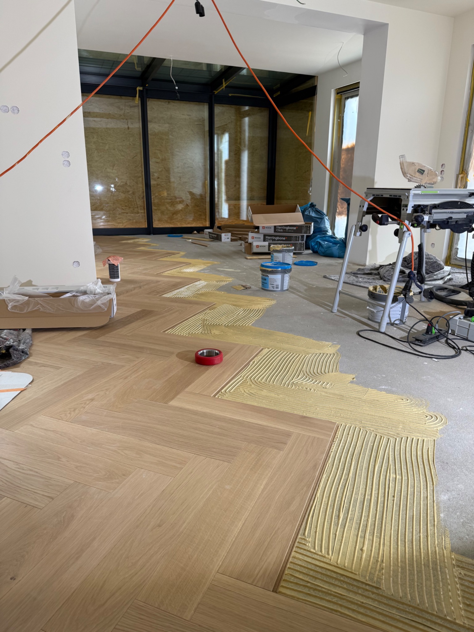 Układanie parkietu w jodełkę: widoczny klej i fragment ułożonej podłogi. W tle narzędzia i materiały budowlane. Jasne wnętrze z dużymi oknami.