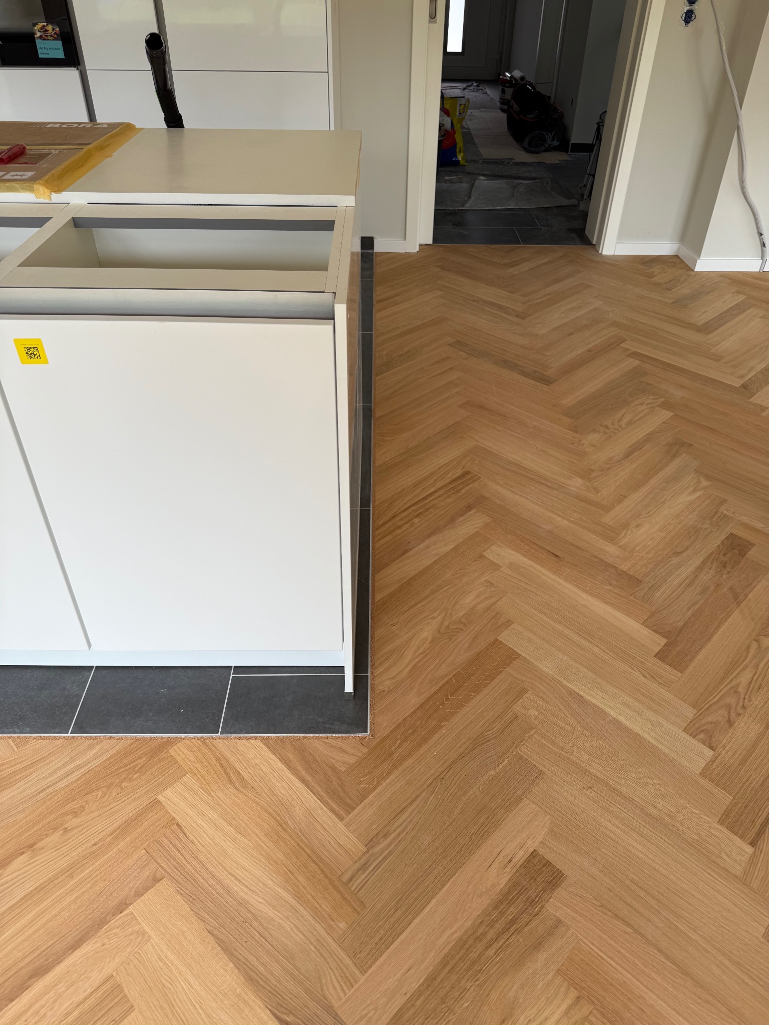 Parkiet ułożony w jodełkę francuską, z widoczną białą zabudową kuchenną i fragmentem korytarza z ciemnymi płytkami. Jasne drewno parkietu.