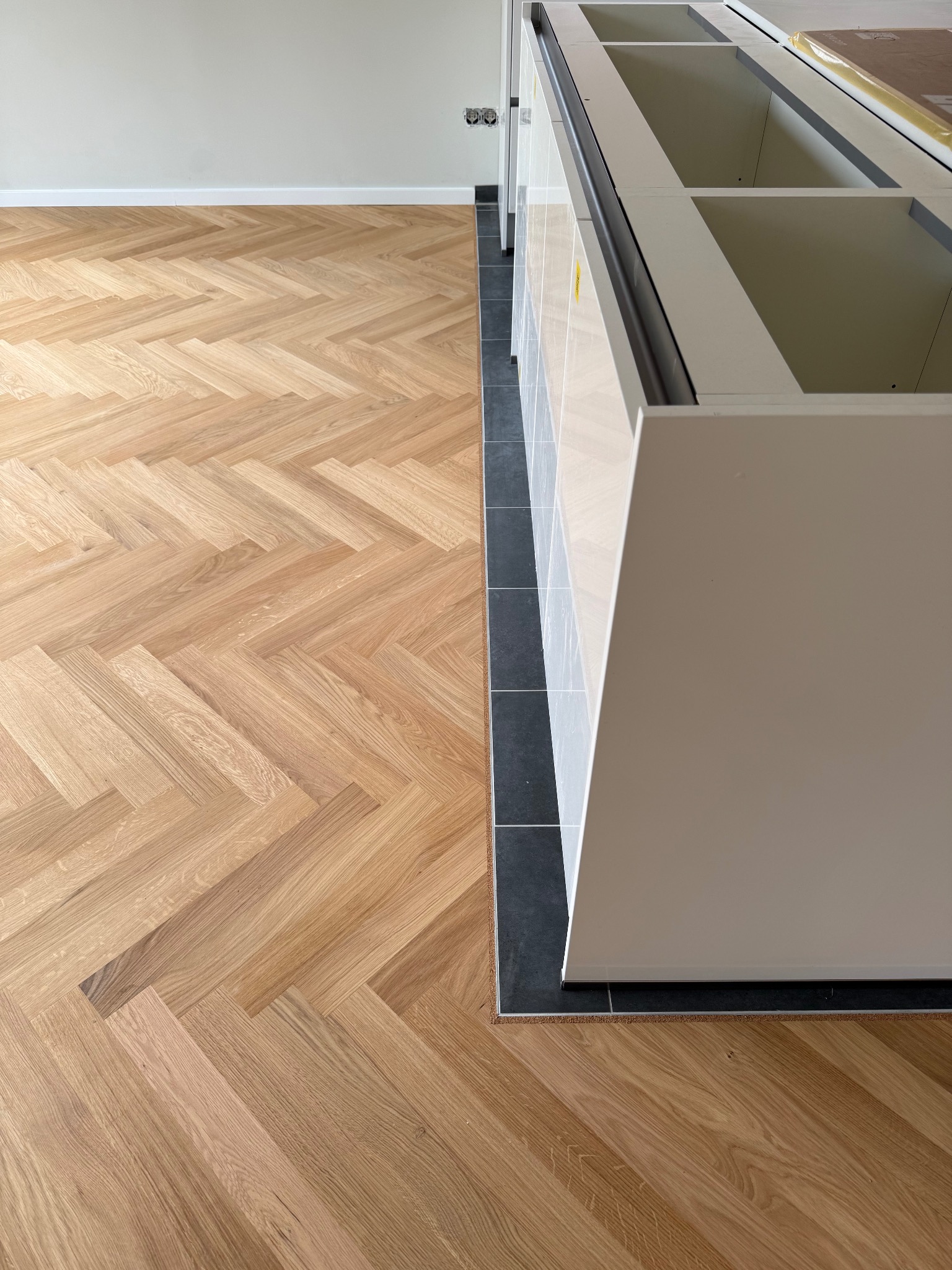 Ułożony parkiet w jodełkę przy zabudowie kuchennej z czarnymi płytkami podłogowymi. Jasne drewno parkietu kontrastuje z ciemnym kolorem płytek.