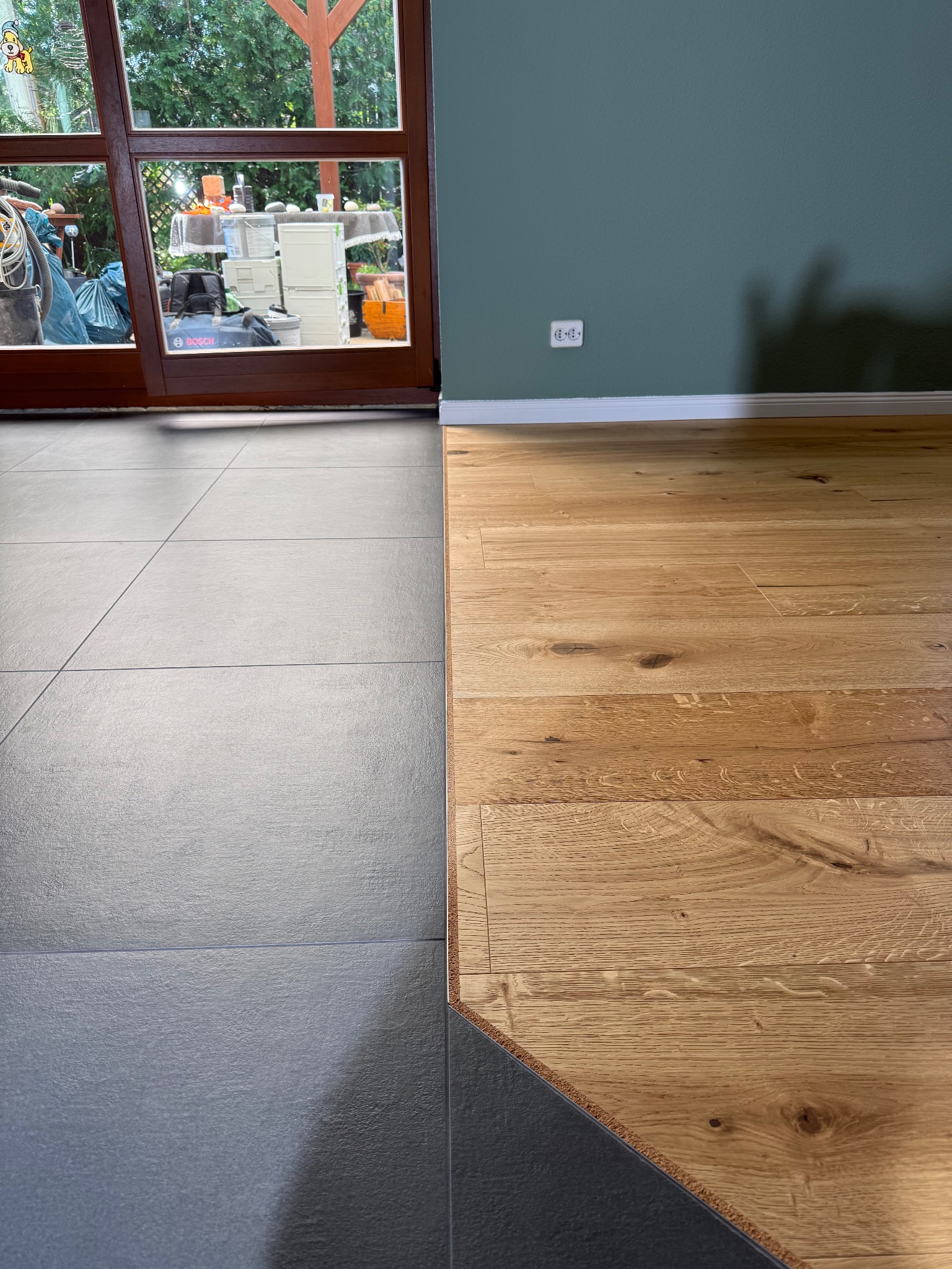 Połączenie szarej płytki z jasnym drewnianym parkietem w nowym wnętrzu. Widoczne listwy przypodłogowe i okno z widokiem na zieleń.