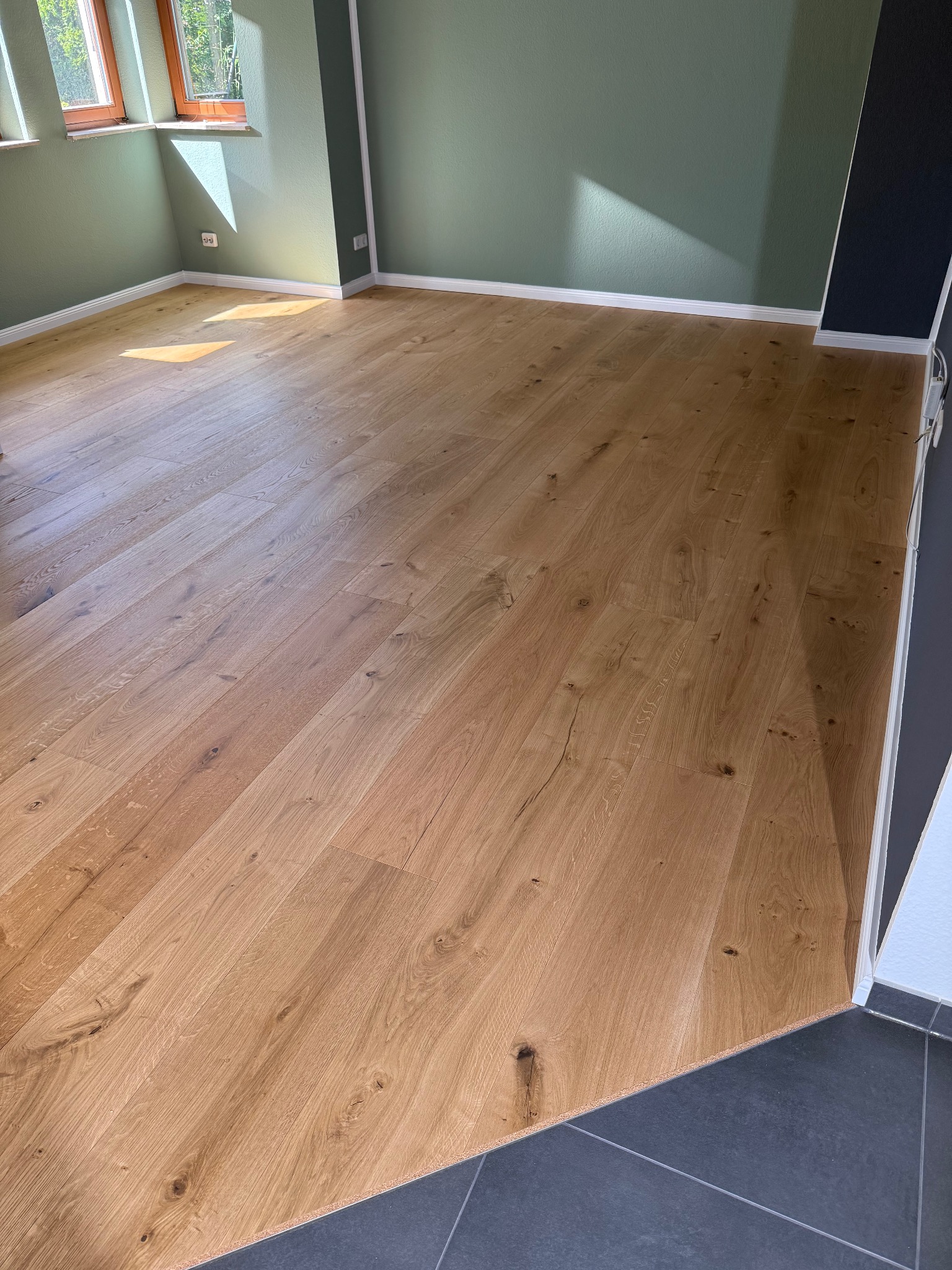 Jasny, dębowy parkiet ułożony w nowym wnętrzu, kontrastujący z ciemnymi płytkami w przejściu. Świeże, zielone ściany i naturalne światło podkreślają fakturę drewna.