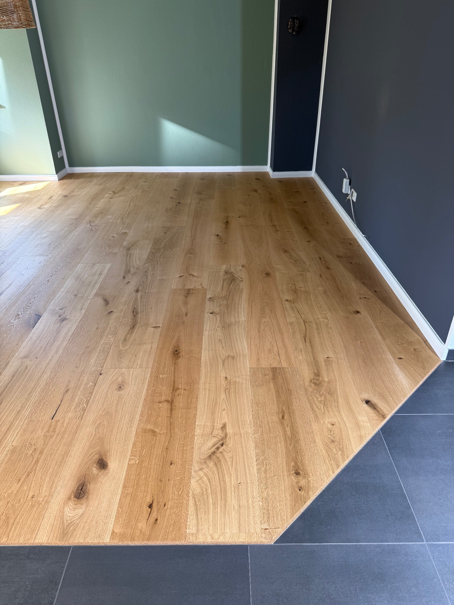 Jasny, dębowy parkiet ułożony w pokoju z zielono-granatowymi ścianami. Fragment podłogi wykończony szarymi płytkami. Widoczne detale drewna i minimalistyczny design.