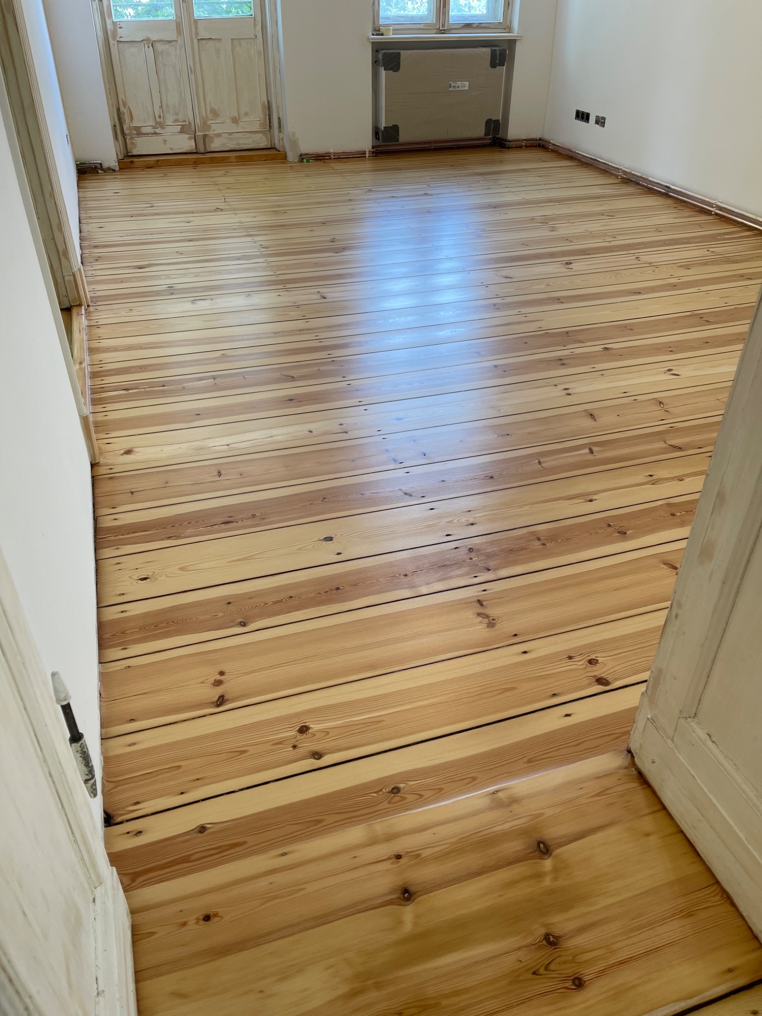 Jasne, odnowione deski podłogowe z widocznymi słojami drewna, w tle białe, odrapane drzwi i grzejnik obudowany drewnem.