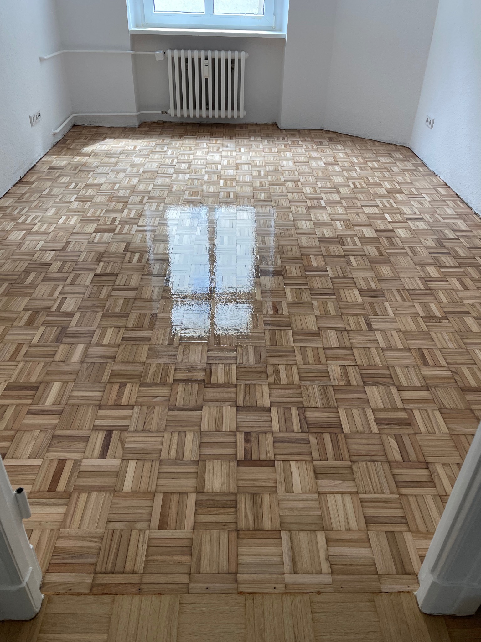 Parkiet ułożony w kwadraty, po zabiegu odświeżającym, z widocznym odbiciem okna, w pustym pokoju z białymi ścianami i grzejnikiem.