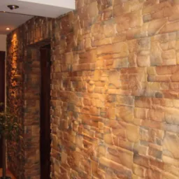 Ściana wykończona dekoracyjnym kamieniem w odcieniach beżu i brązu, oświetlona ciepłym światłem, z widocznym fragmentem drewnianych drzwi i rośliną doniczkową w tle.