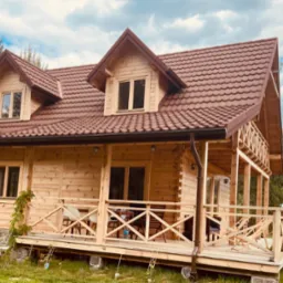 Nowoczesny dom z bali z brązowym dachem i zadaszonym tarasem, widok z zewnątrz w słoneczny dzień.