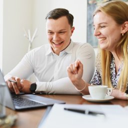 Kamil Tomyślak Nieruchomości - Uśmiechnięci konsultanci analizują ofertę na laptopie, filiżanki kawy na stole, jasne wnętrze biura.