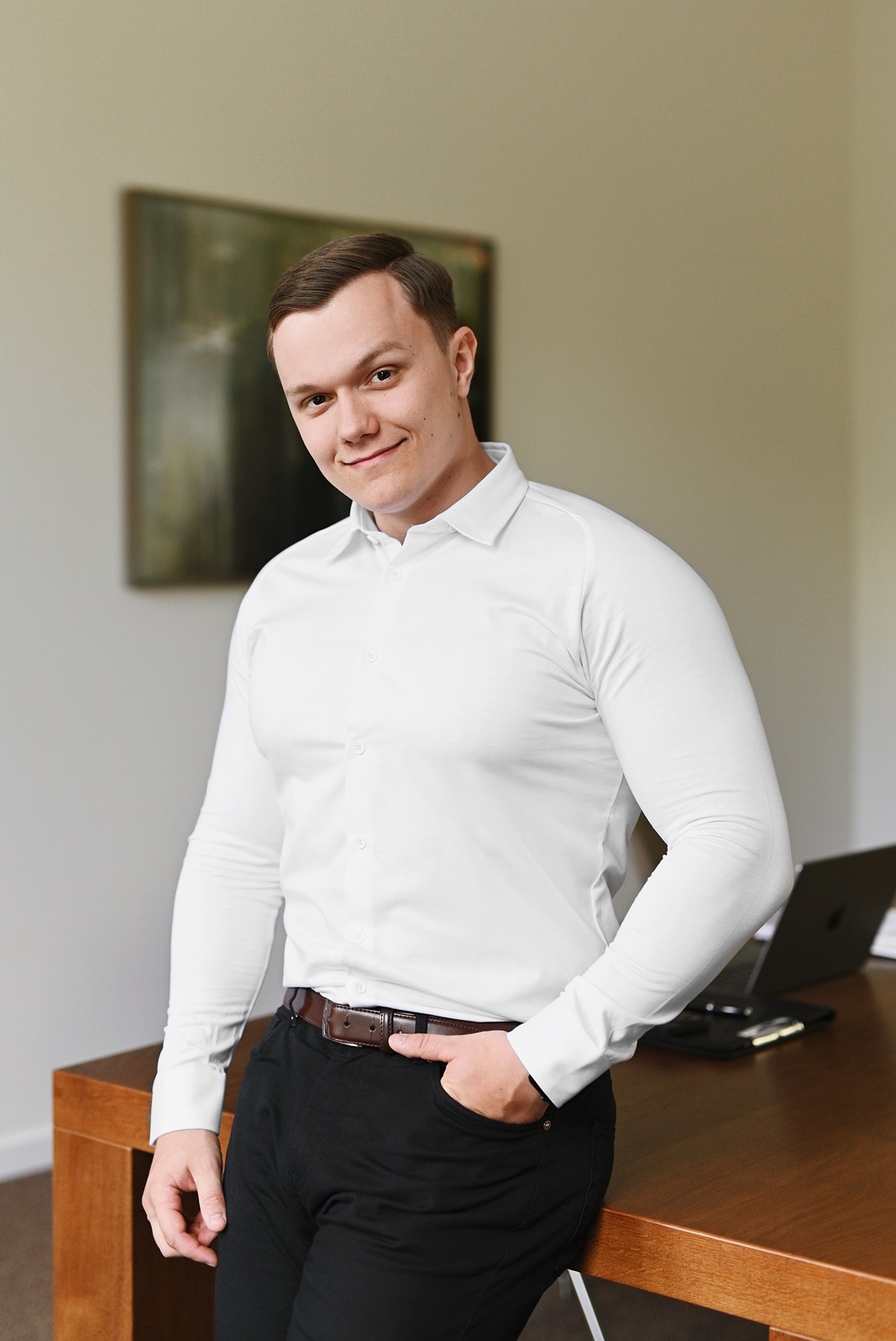 Młody agent nieruchomości w białej koszuli, opierający się o drewniane biurko z laptopem w tle, prezentujący profesjonalny wizerunek.