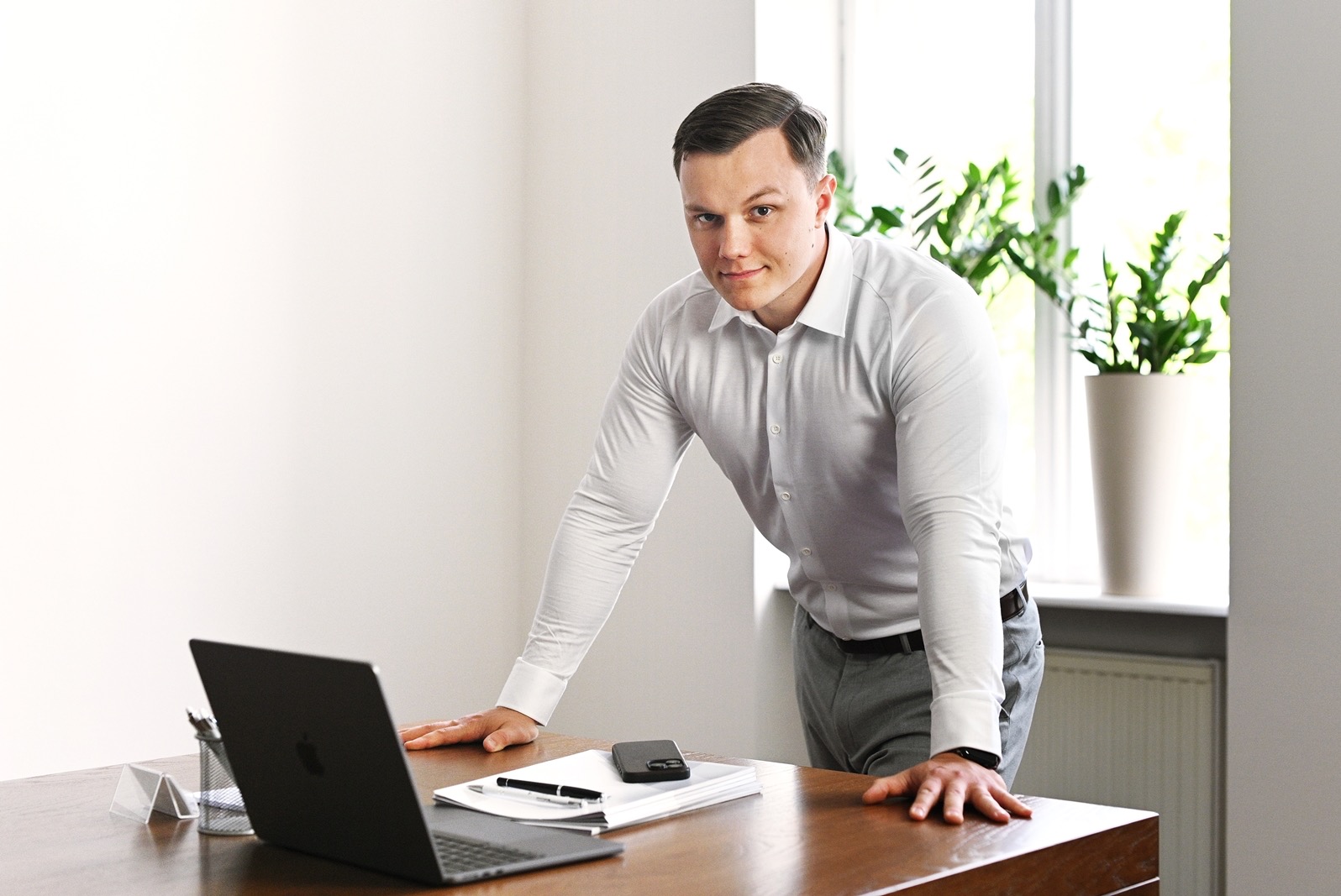 Młody mężczyzna w jasnej koszuli opiera się o biurko z laptopem, notatnikiem i telefonem, w tle okno z rośliną doniczkową.