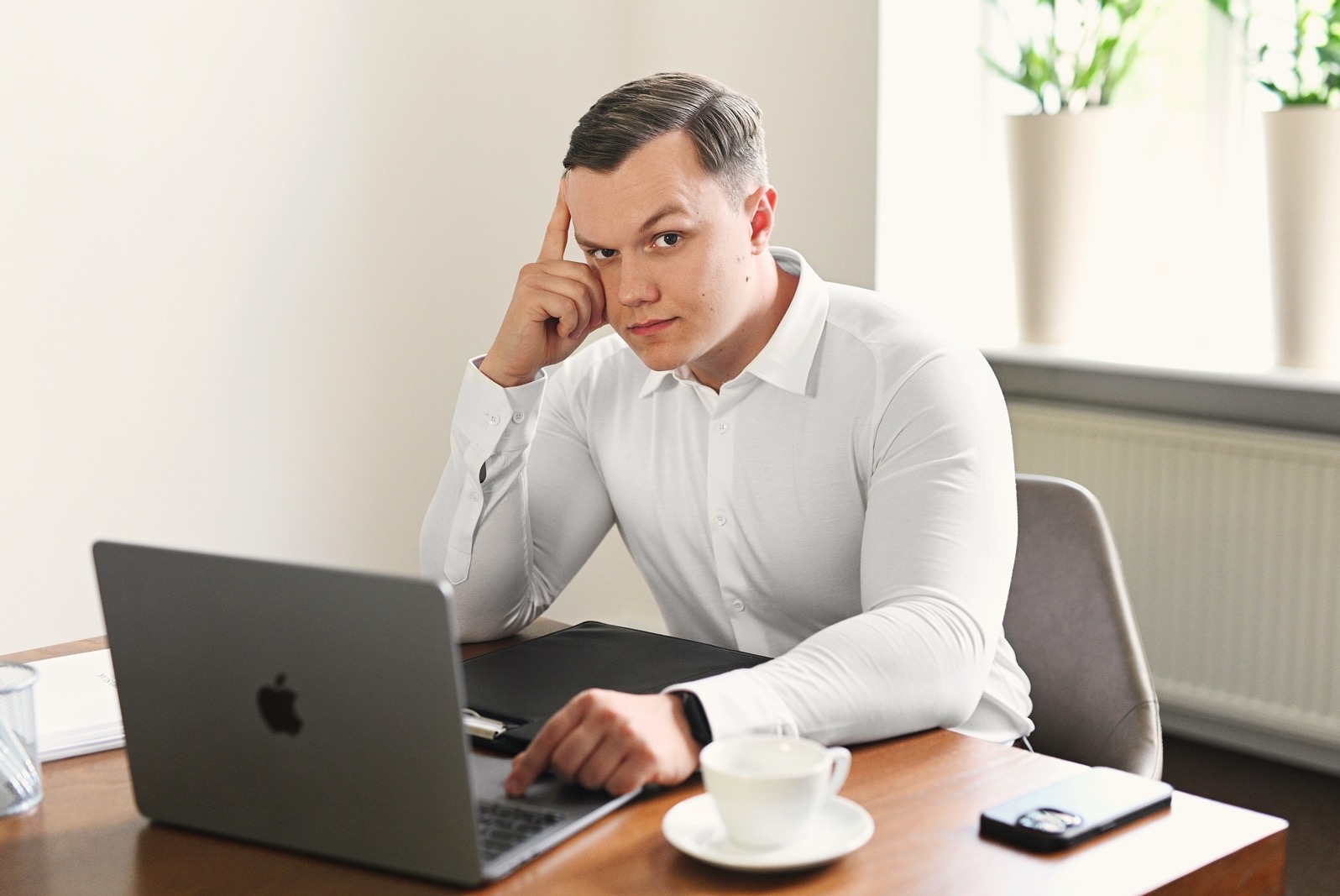 Mężczyzna w białej koszuli siedzi przy biurku z laptopem i filiżanką, zamyślony, dotykając czoła palcem, w tle okno z roślinami doniczkowymi.