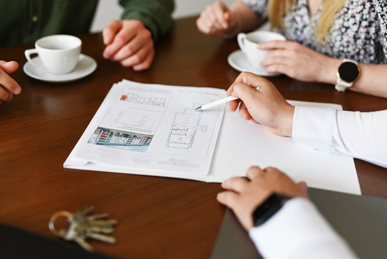Spotkanie przy drewnianym stole: osoba wskazująca długopisem na plan mieszkania, obok klucze, filiżanki i dłonie uczestników.