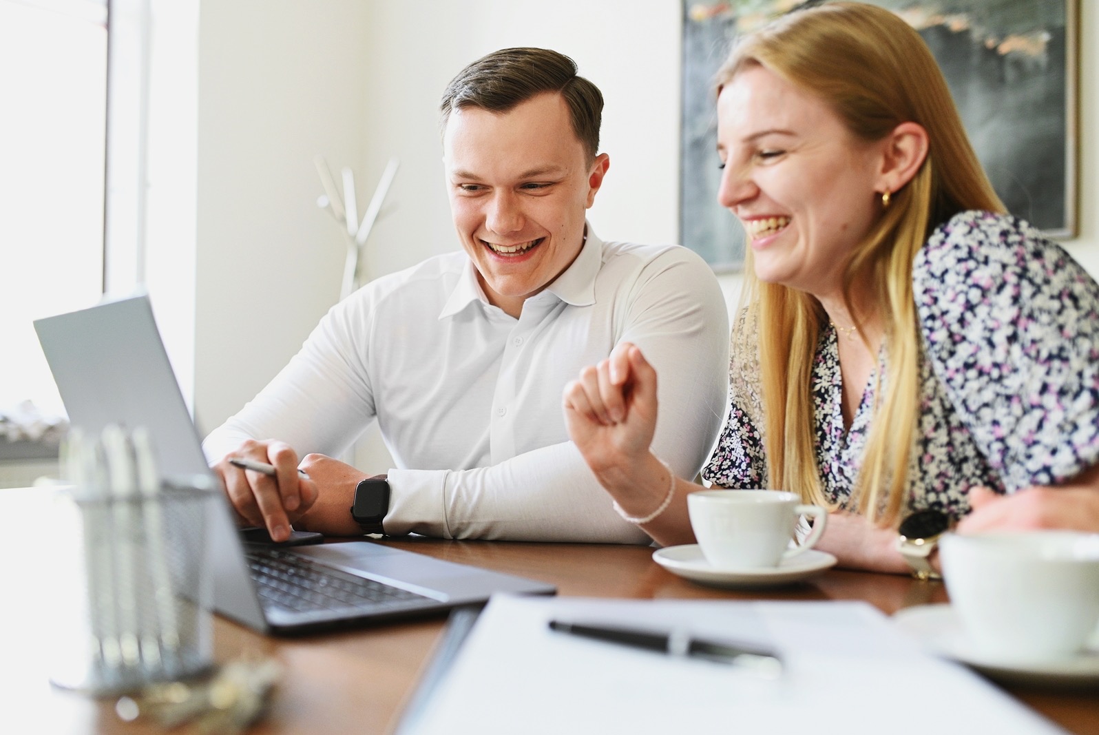 Uśmiechnięci konsultanci analizują ofertę na laptopie, filiżanki kawy na stole, jasne wnętrze biura.
