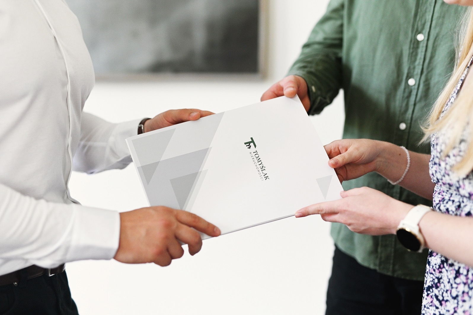 Wręczenie dokumentu z logo agencji nieruchomości Tomyslak pomiędzy trzema osobami, z widocznymi dłońmi i fragmentami ubrań, na jasnym tle.