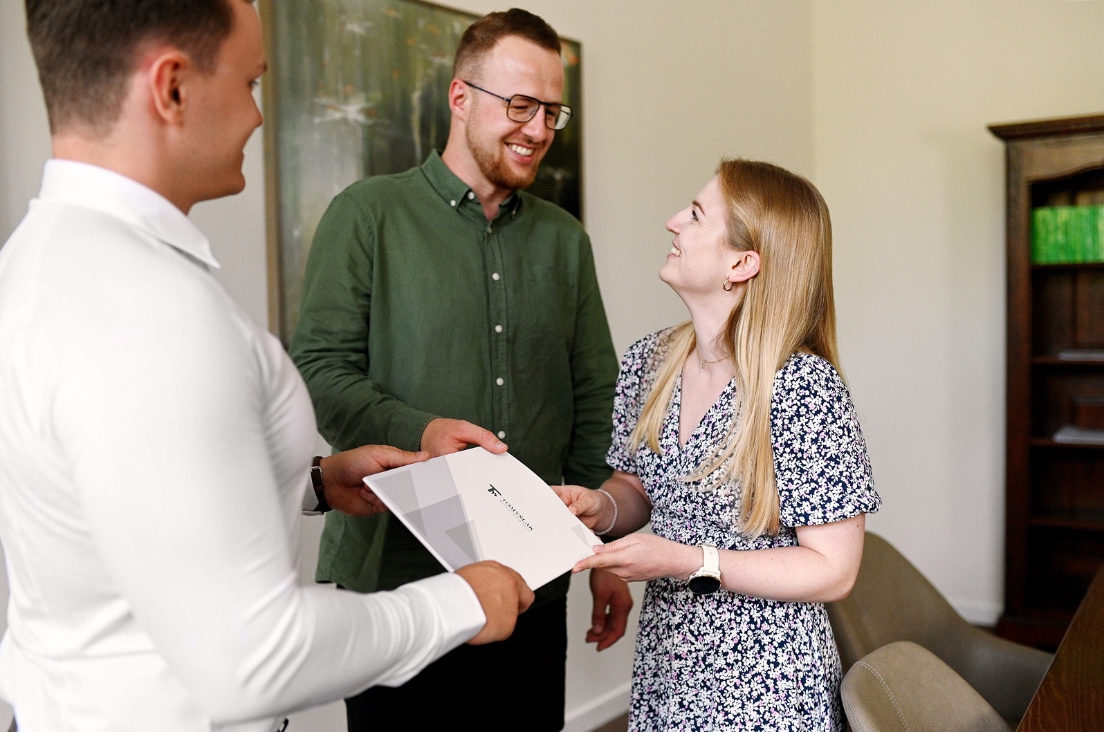 Młoda kobieta w kwiecistej sukience odbiera dokumenty od agenta nieruchomości, uśmiechając się do mężczyzny w zielonej koszuli, na ścianie wisi abstrakcyjny obraz.