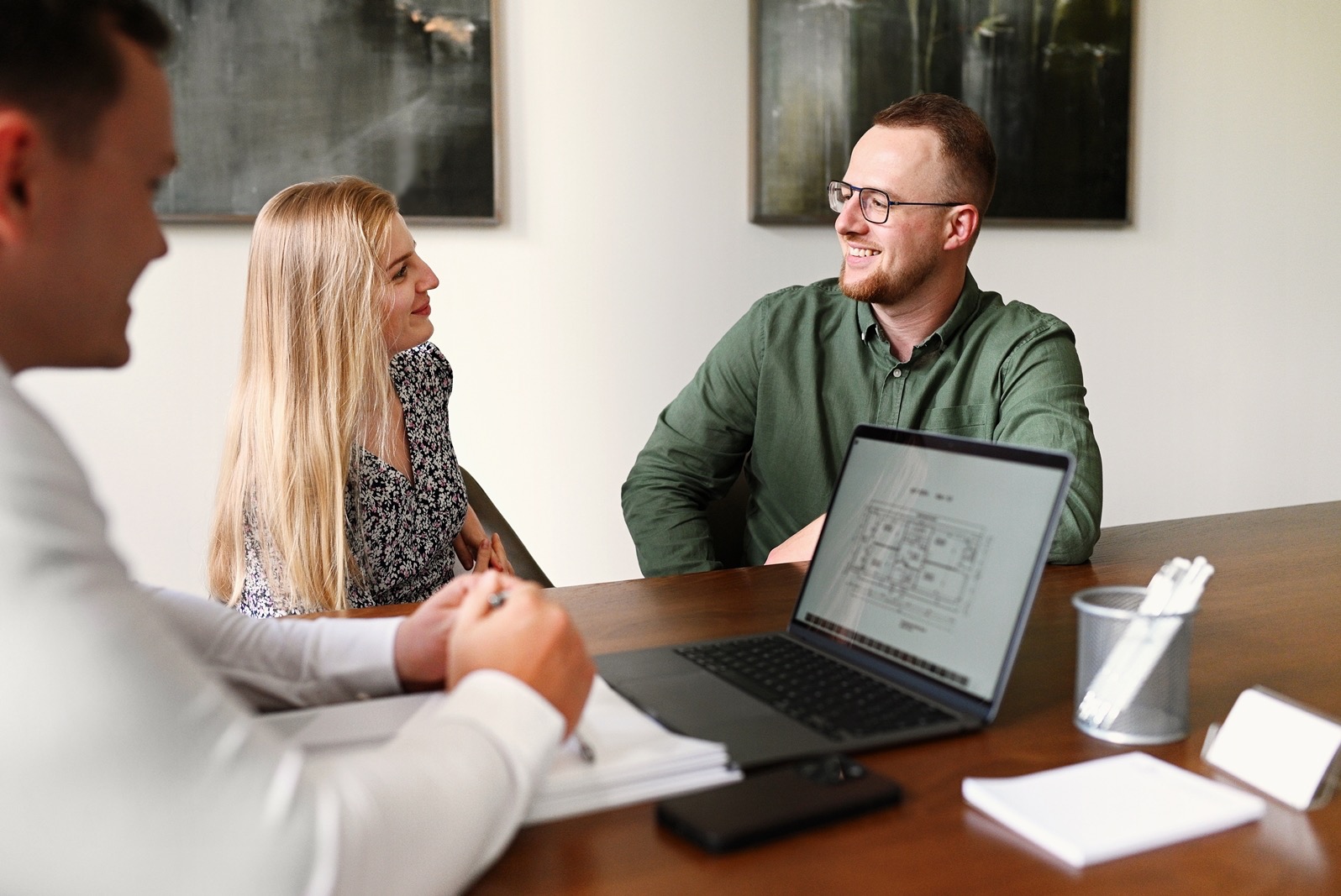 Para omawia projekt mieszkania na laptopie z agentem nieruchomości przy drewnianym stole, widoczne szkice planu, jasne ściany i obrazy w tle.