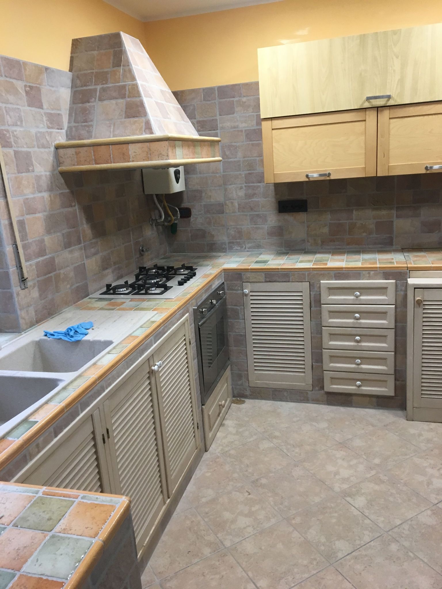 Jasna kuchnia z terakotową mozaiką na ścianach i blacie, drewnianymi szafkami z żaluzjowymi drzwiczkami, widocznym zlewem z niebieską ścierką i kuchenką gazową.