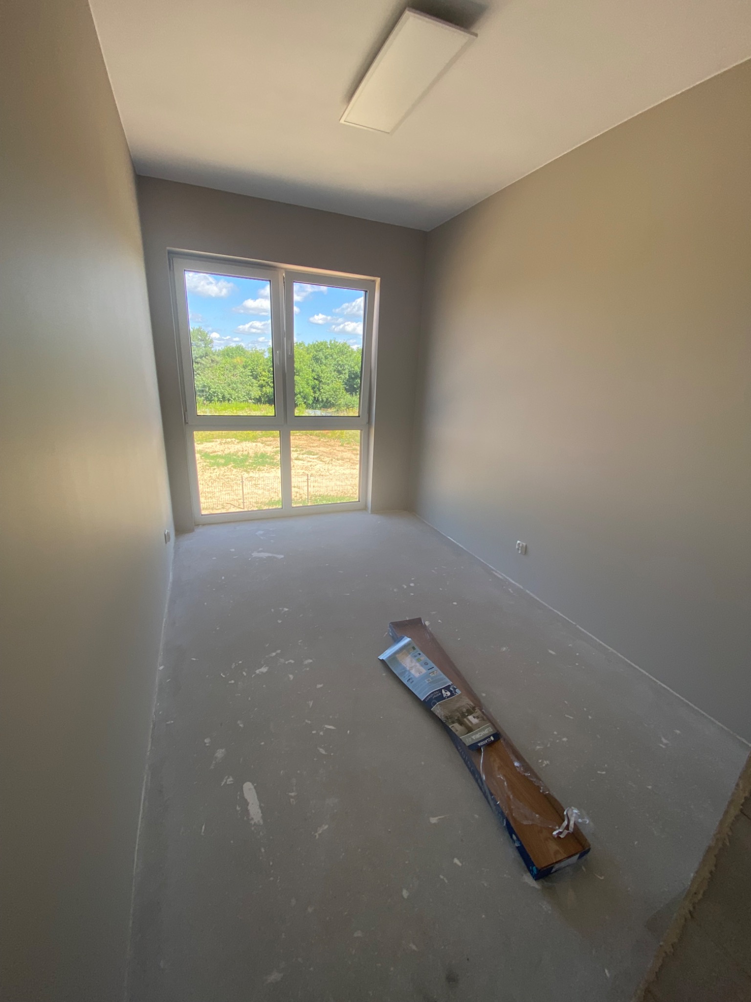 Przygotowanie do ułożenia paneli podłogowych w pustym, szarym pokoju z widokiem na zieleń za oknem; paczka paneli leży na surowej podłodze.