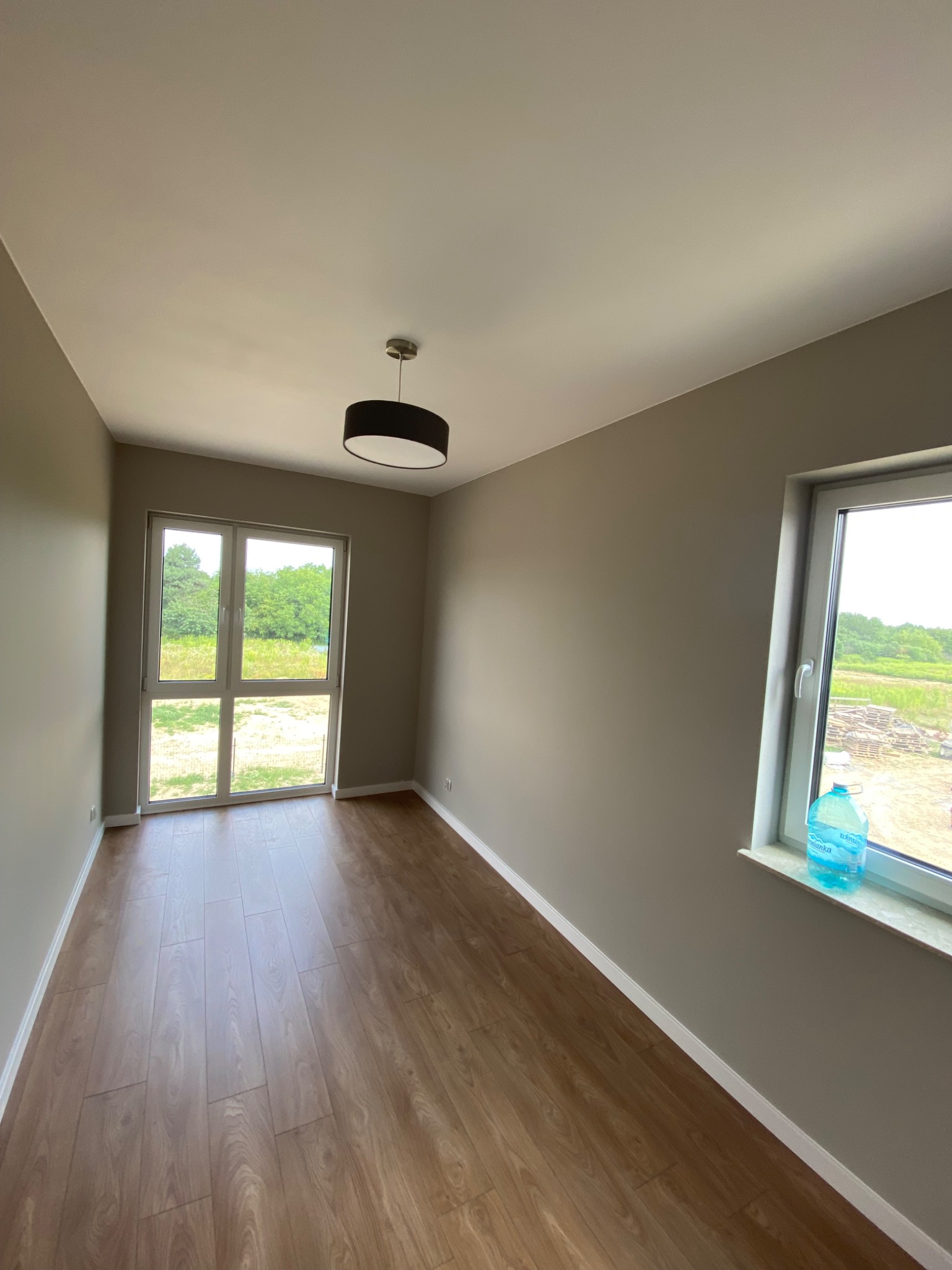 Pomalowane na szaro puste pomieszczenie z panelami podłogowymi, czarną lampą sufitową i butelką wody na parapecie okiennym.