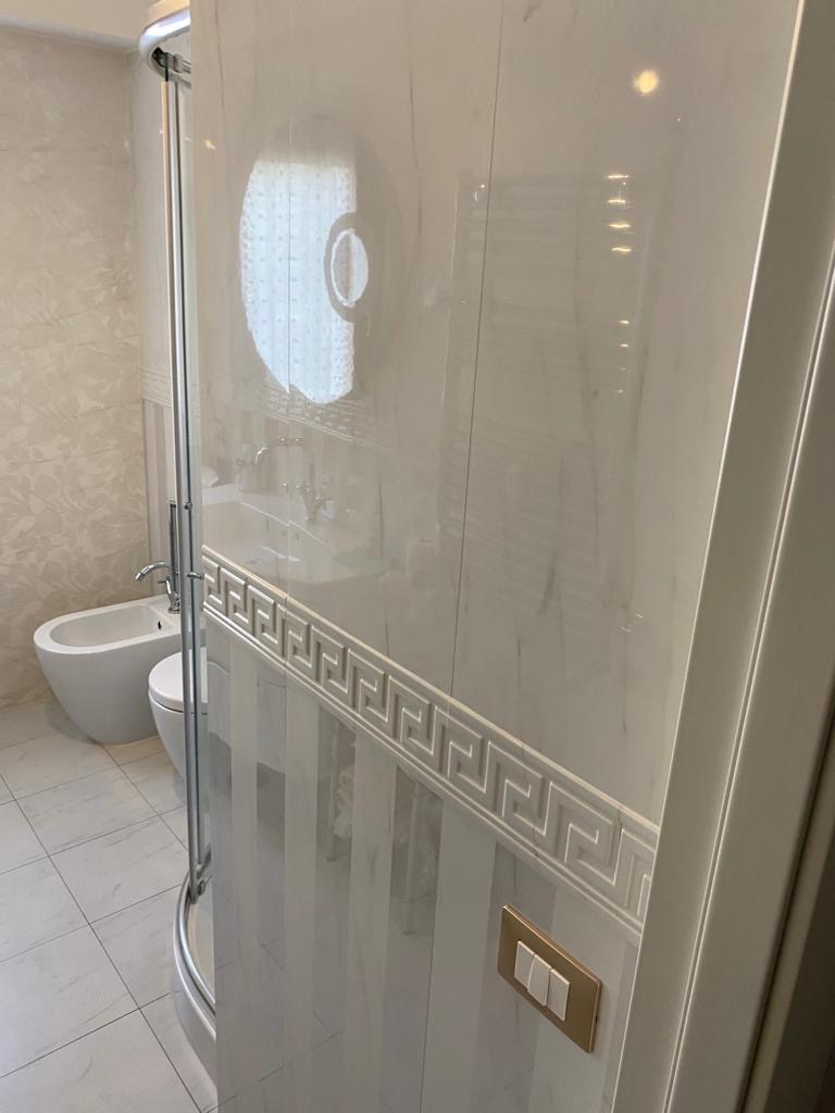 Wykończona łazienka z kabiną prysznicową, bidetem i toaletą. Białe płytki z dekoracyjnym wzorem meandry na ścianie. Złoty włącznik światła na ścianie.