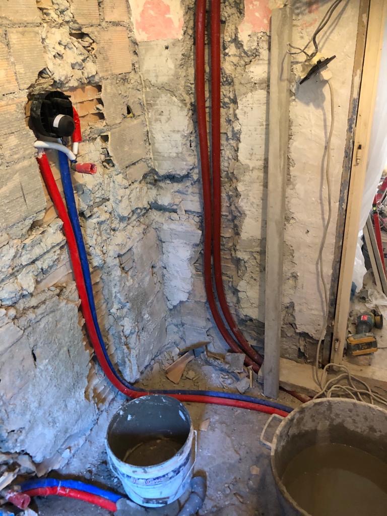 Surowa ściana z odsłoniętą cegłą, nowe rury wodne w kolorze czerwonym i niebieskim, puszka instalacyjna, dwa wiadra z zaprawą, widoczne przewody elektryczne i resztki starej instalacji.