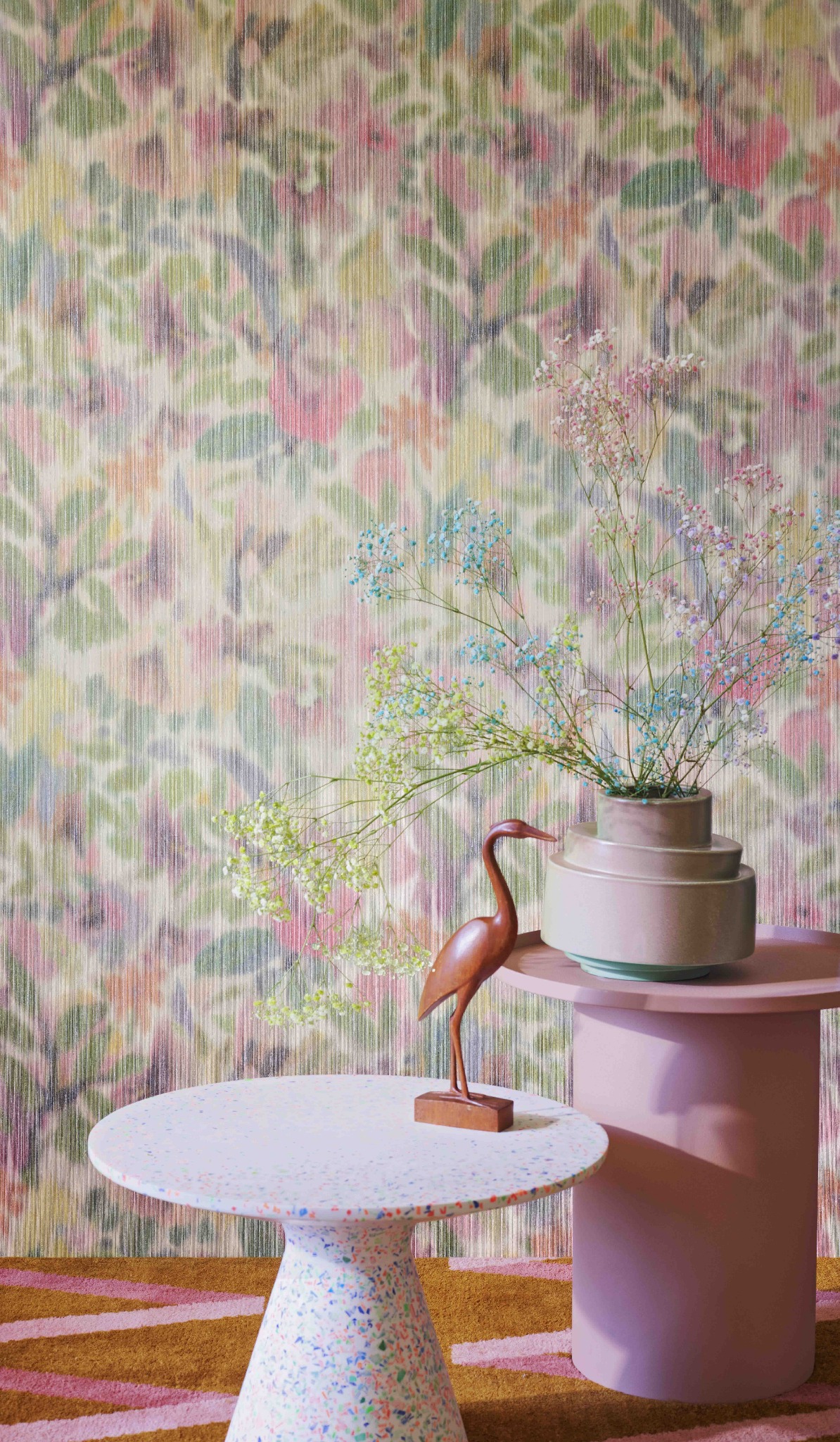 Dwa stoliki kawowe: okrągły, biały z kolorowymi plamkami i różowy, cylindryczny, na tle tapety w kwiaty i dywanu w pasy.