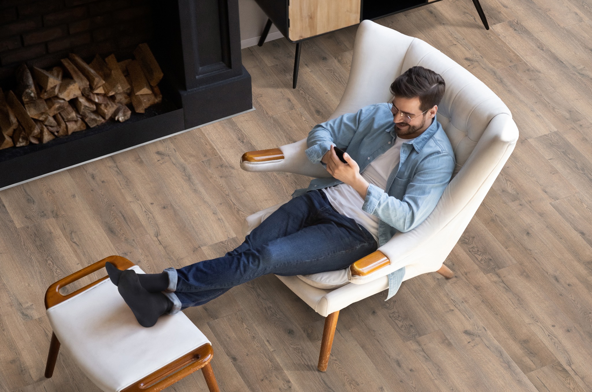 Mężczyzna relaksuje się w fotelu, przeglądając telefon na nowej, jasnej podłodze z paneli drewnianych, obok kominka z ułożonym drewnem.