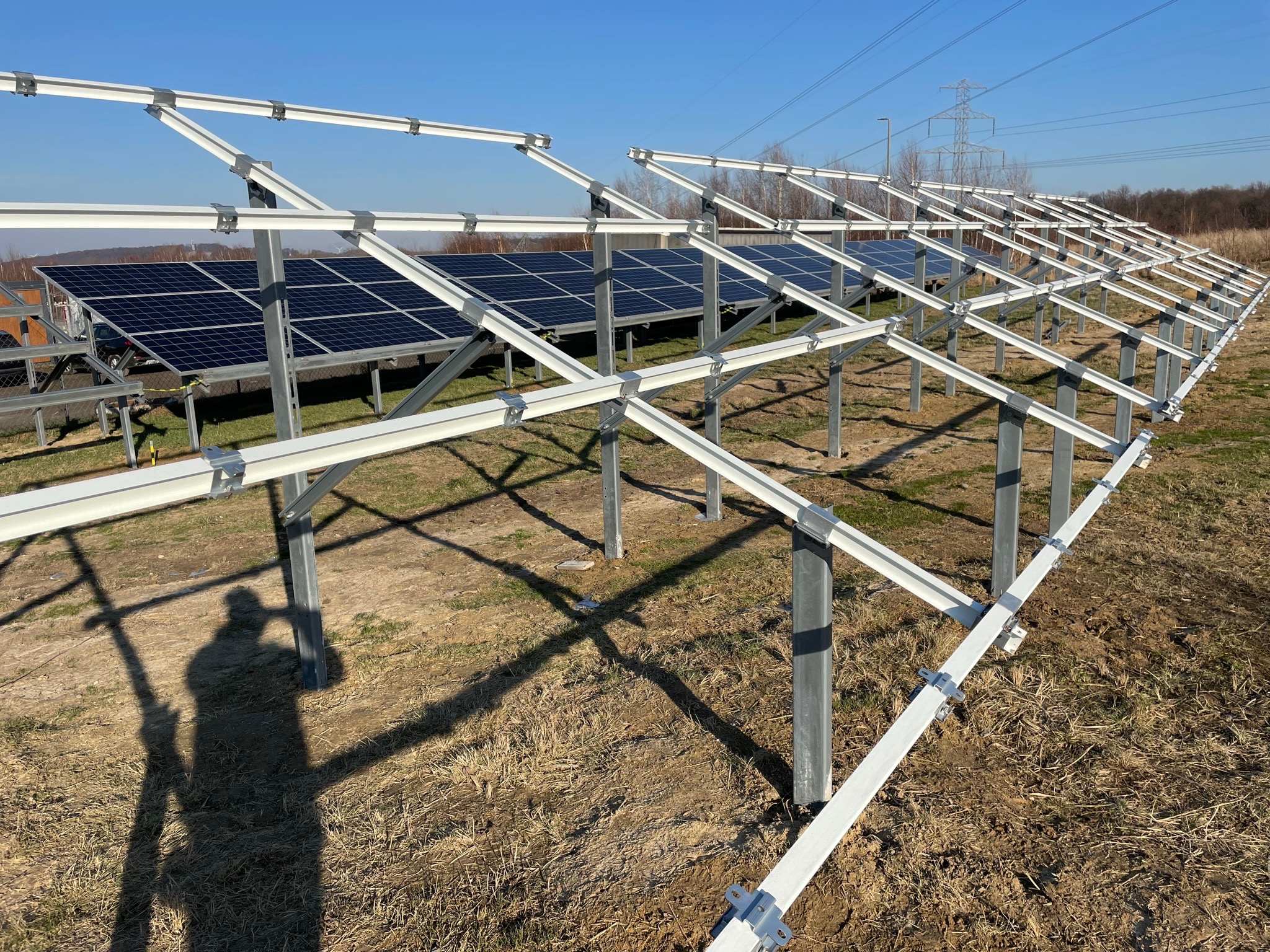 Montaż paneli fotowoltaicznych na konstrukcji wsporczej, widoczny cień osoby fotografującej na trawie.