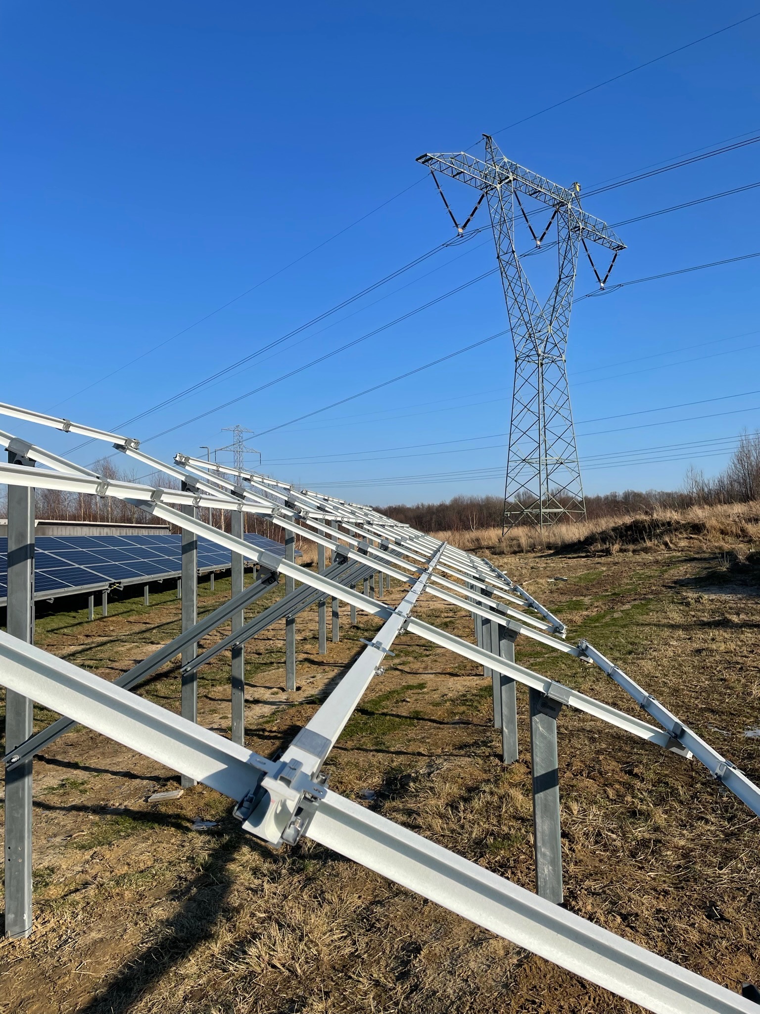 Konstrukcja wsporcza pod panele fotowoltaiczne w trakcie montażu, widoczna linia wysokiego napięcia i częściowo zamontowane panele w tle, słoneczny dzień.