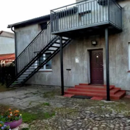 Zewnętrzne schody metalowe z balkonem, prowadzące do drzwi wejściowych budynku o szarej elewacji. Na pierwszym planie brukowane podwórze i donice z kwiatami.