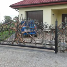 Kuta brama wjazdowa i furtka z motywem drzewa, czarne elementy konstrukcyjne, jasny dom w tle.