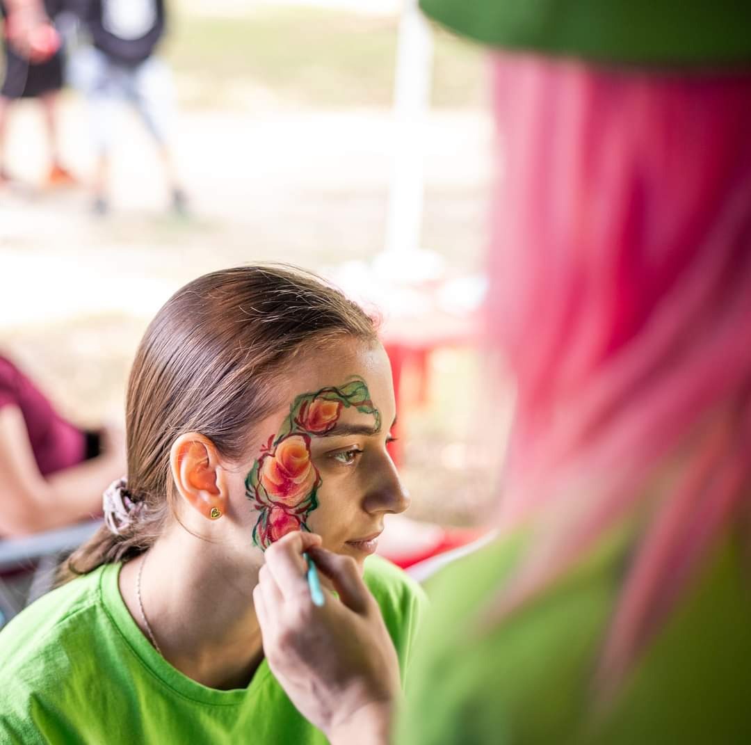 Młoda kobieta z malunkiem kwiatowym na twarzy, wykonywanym przez osobę z różowymi włosami, w plenerze.