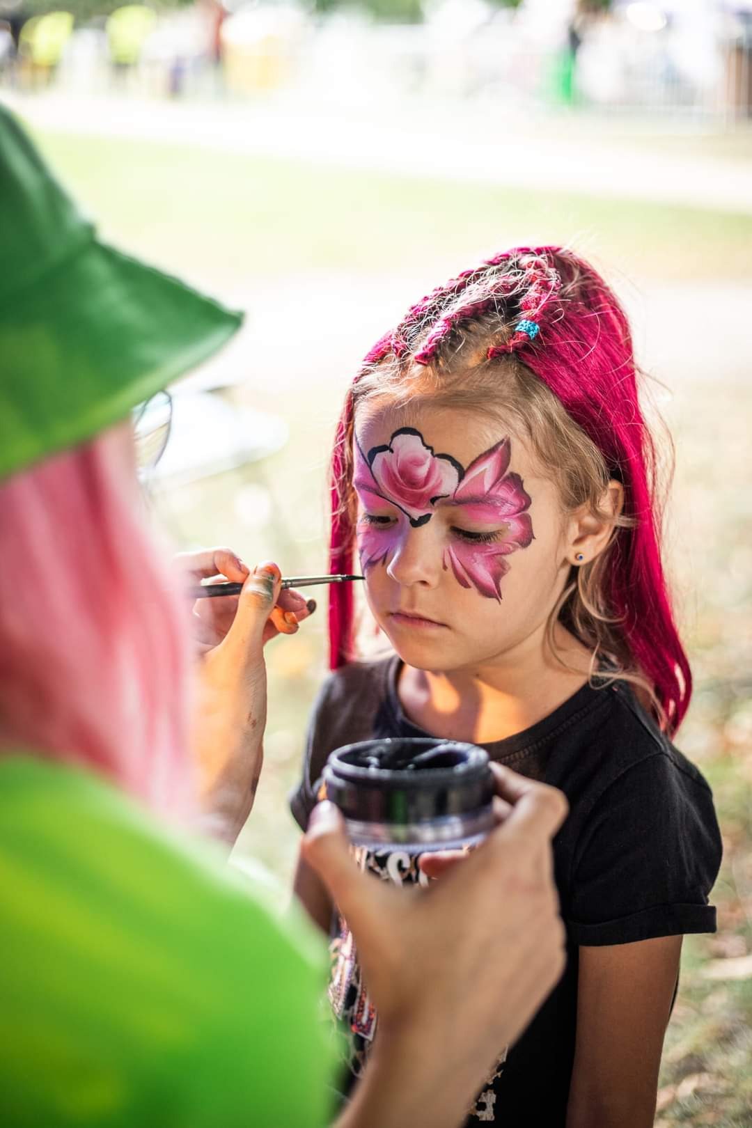 Animator maluje twarz dziewczynki w motyw kwiatowy, używając pędzla i czarnej farby. Dziecko ma różowe włosy z wplecionymi warkoczykami i niebieskimi koralikami.