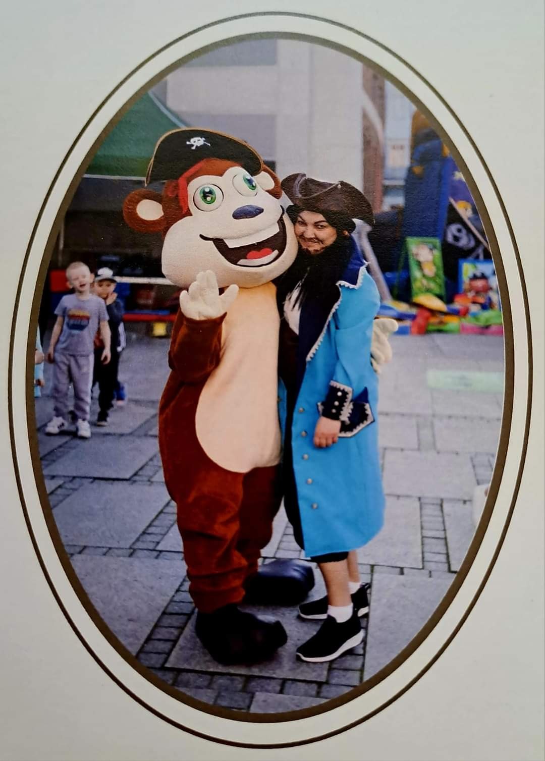 Uśmiechnięta kobieta w kostiumie pirata obejmuje maskotkę małpy-pirata na tle placu zabaw i dzieci.