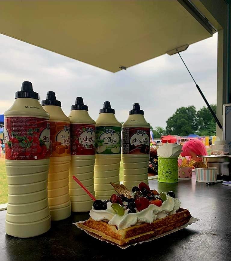 Wafel belgijski z bitą śmietaną i owocami (truskawki, borówki, winogrona) na tle butelek z sosami smakowymi (truskawkowy, toffi, wiśniowy, zielone jabłko, czekoladowy) w punkcie gastronomicznym...