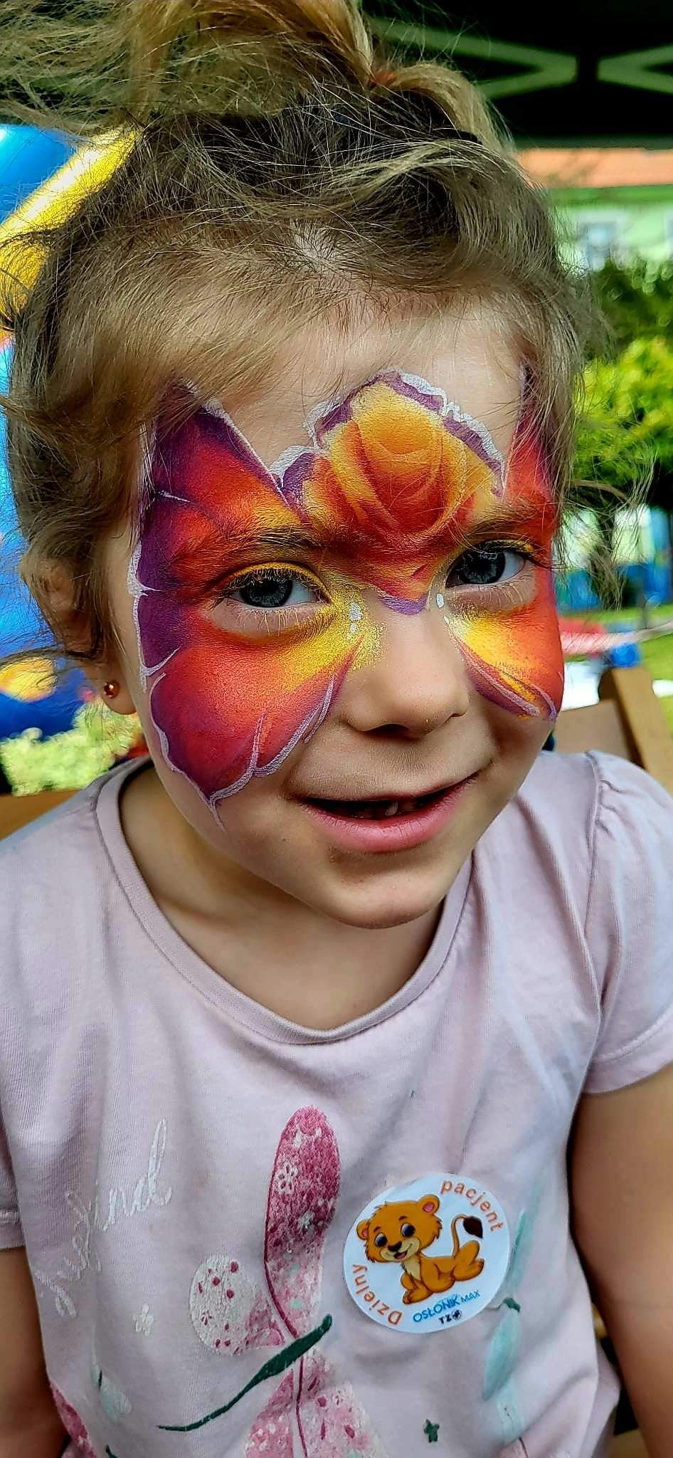 Dziewczynka z pomalowaną twarzą w motywy kwiatowe i motyla, z naklejką 'Dzielny Pacjent' na koszulce, uśmiechnięta.