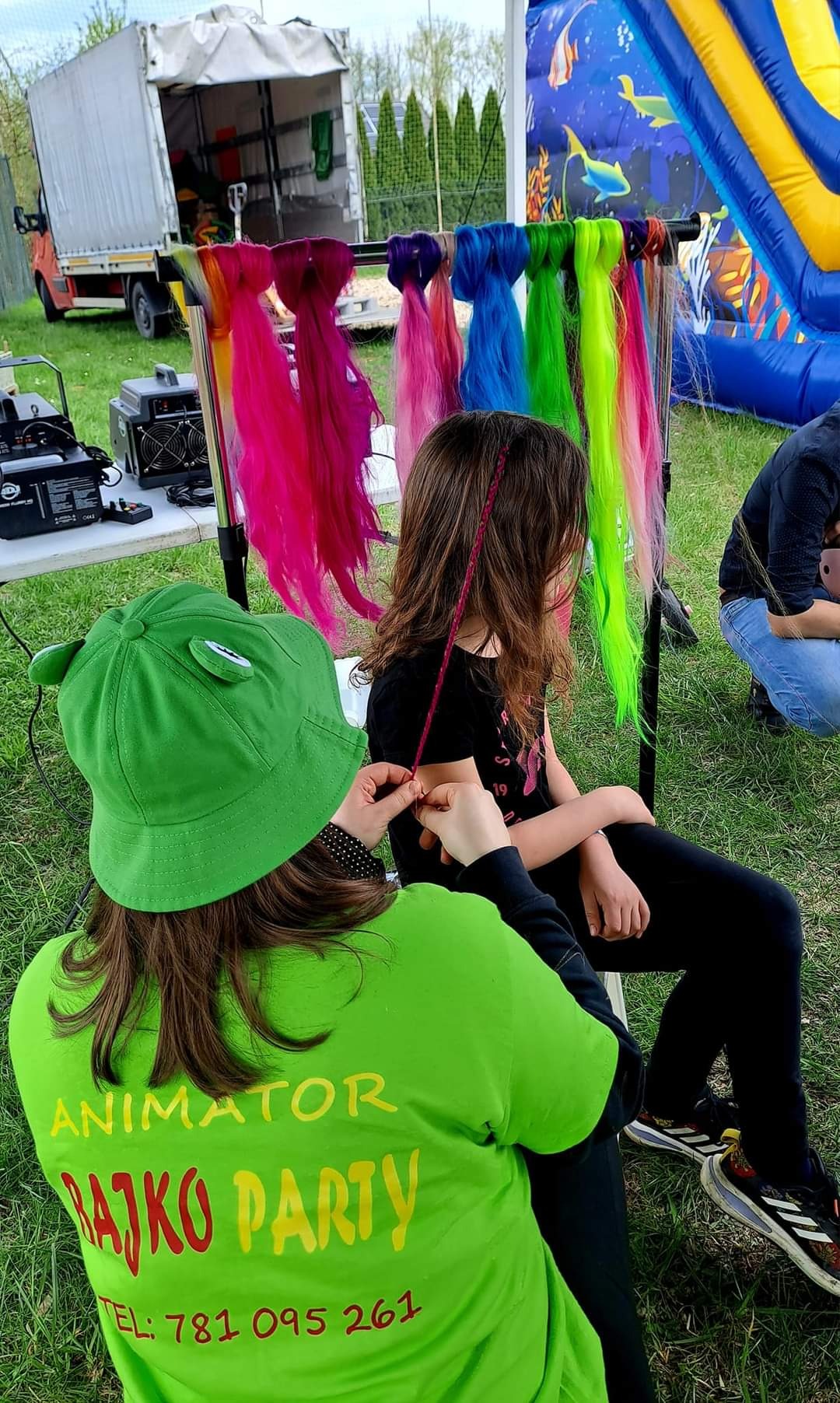 Animator w zielonej koszulce z napisem 'Animator Rajko Party' zaplata kolorowe pasemko włosów dziewczynce siedzącej na krześle, na tle dmuchanego zamku i stojaka z kolorowymi doczepianymi włosami.