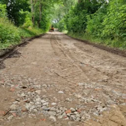 Przygotowanie podłoża drogi gruntowej z gruzem i koleinami, w tle widoczna maszyna budowlana, otoczenie drzew i zieleni.