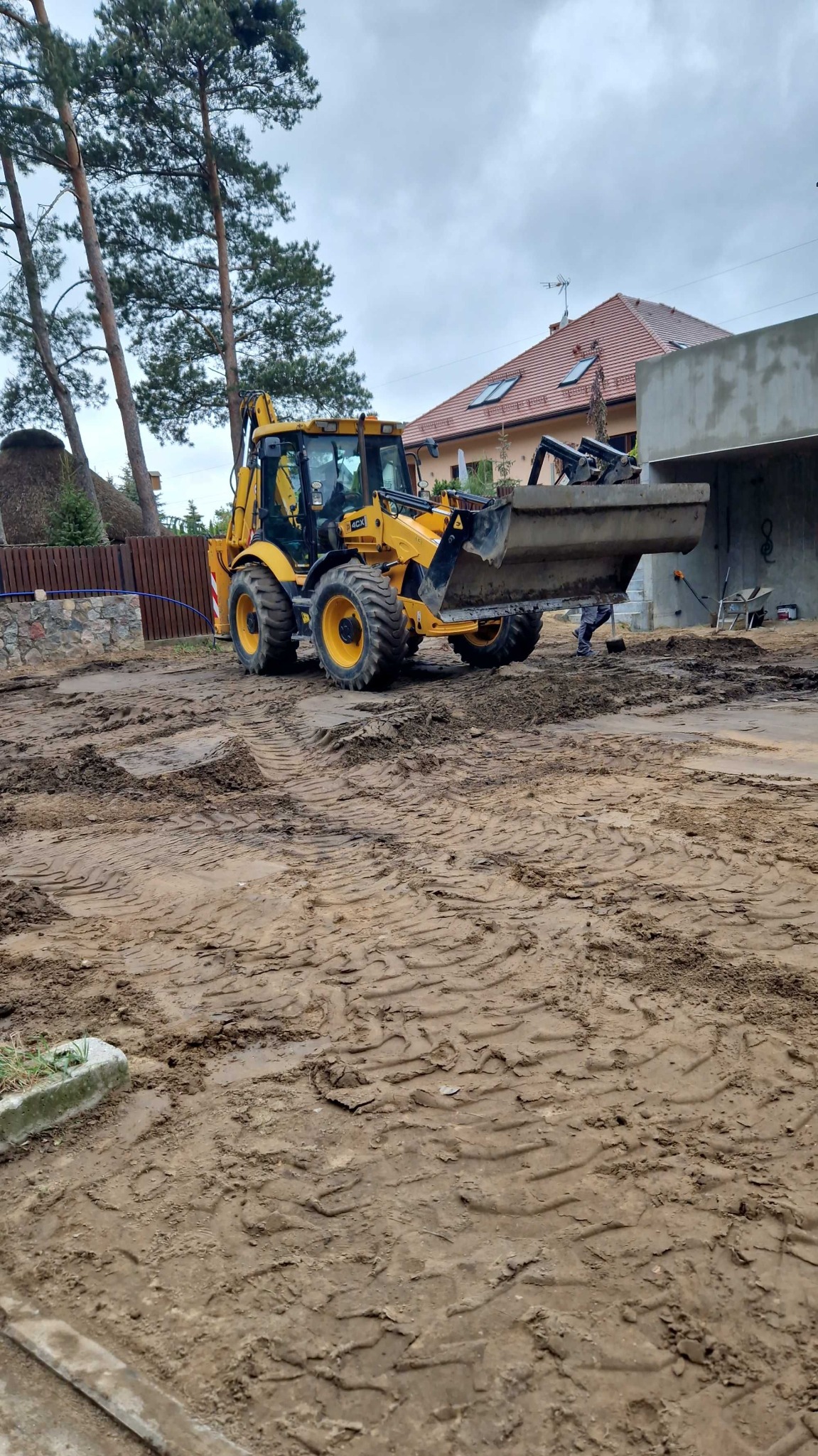 Żółta koparko-ładowarka JCB 4CX na nierównym, błotnistym terenie budowy, widoczne ślady opon, w tle dom jednorodzinny i fragment betonowej konstrukcji.