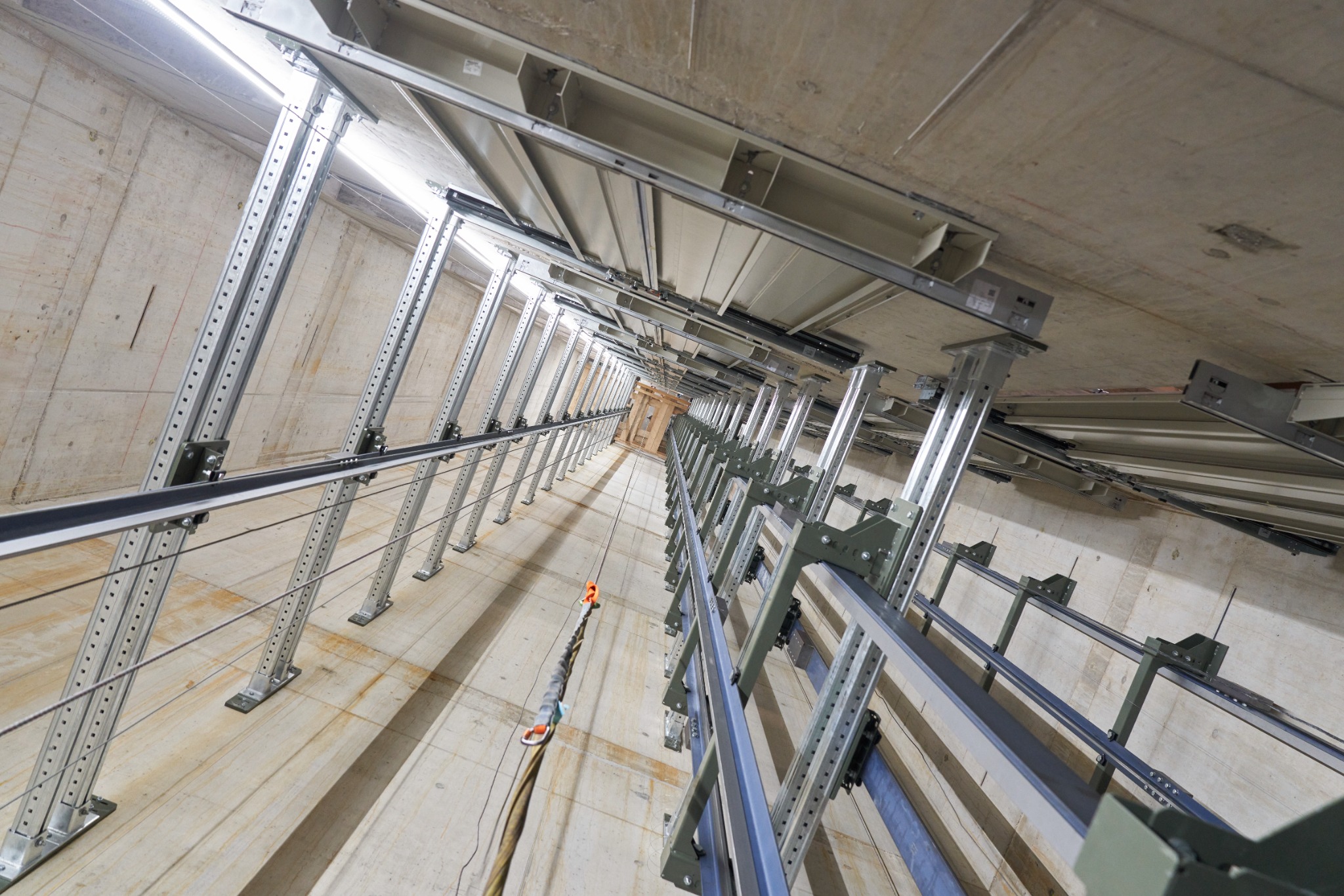 Wnętrze szybu windowego w trakcie montażu, widok z dołu na metalowe prowadnice i konstrukcję nośną, surowy beton na ścianach.