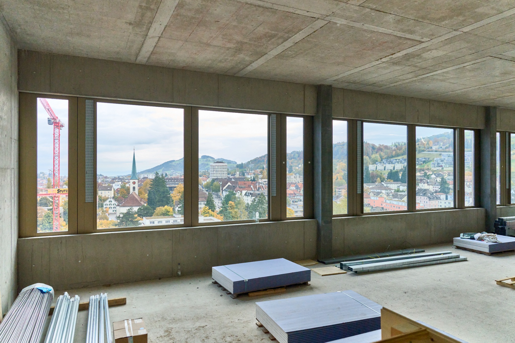 Wnętrze surowego budynku w trakcie budowy, widok przez okna na miasto z kościelną wieżą i dźwigiem.