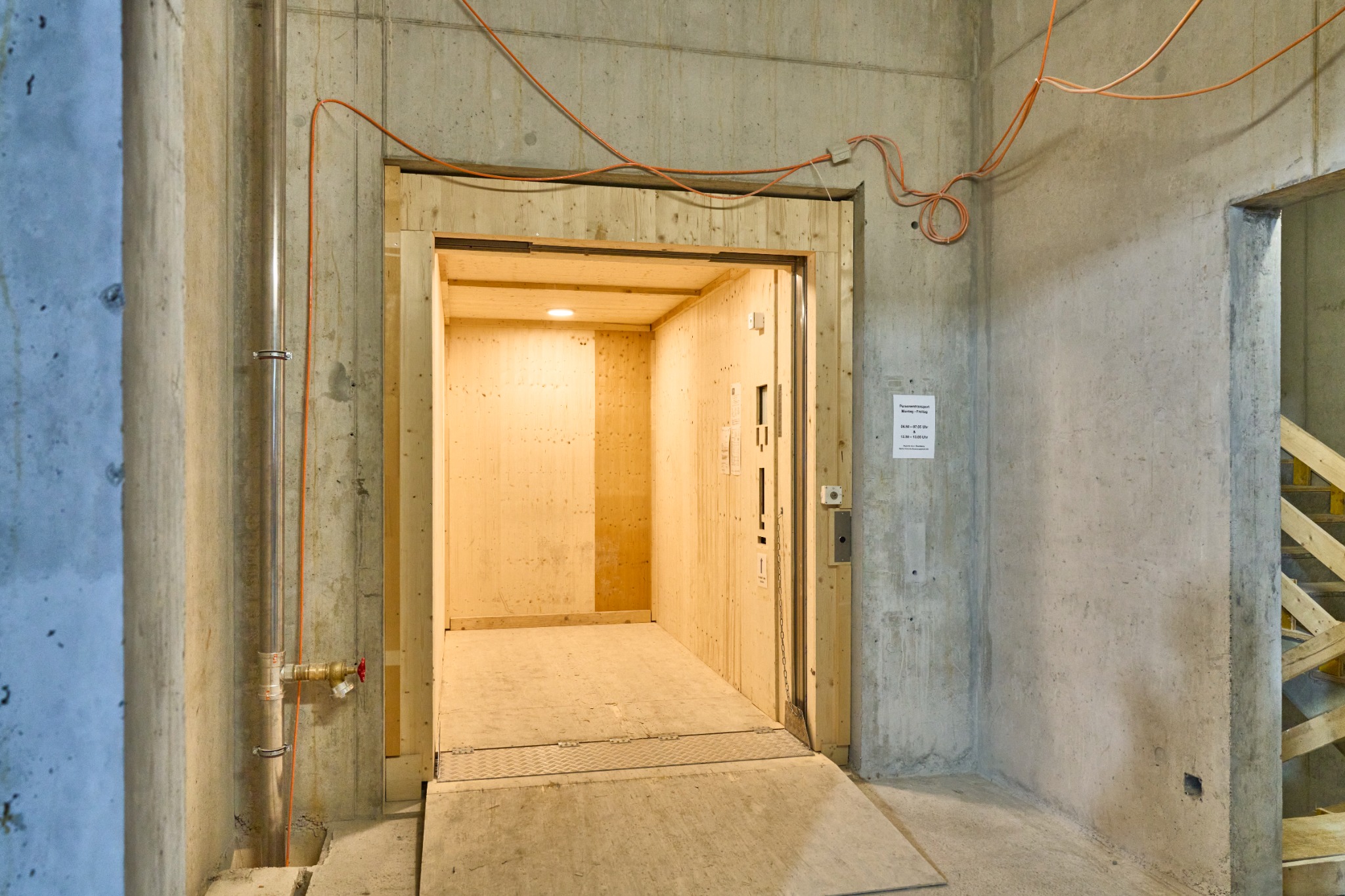 Wnętrze drewnianej kabiny dźwigu towarowego w surowym stanie, betonowe ściany i podłoga, widoczne instalacje elektryczne i rury.