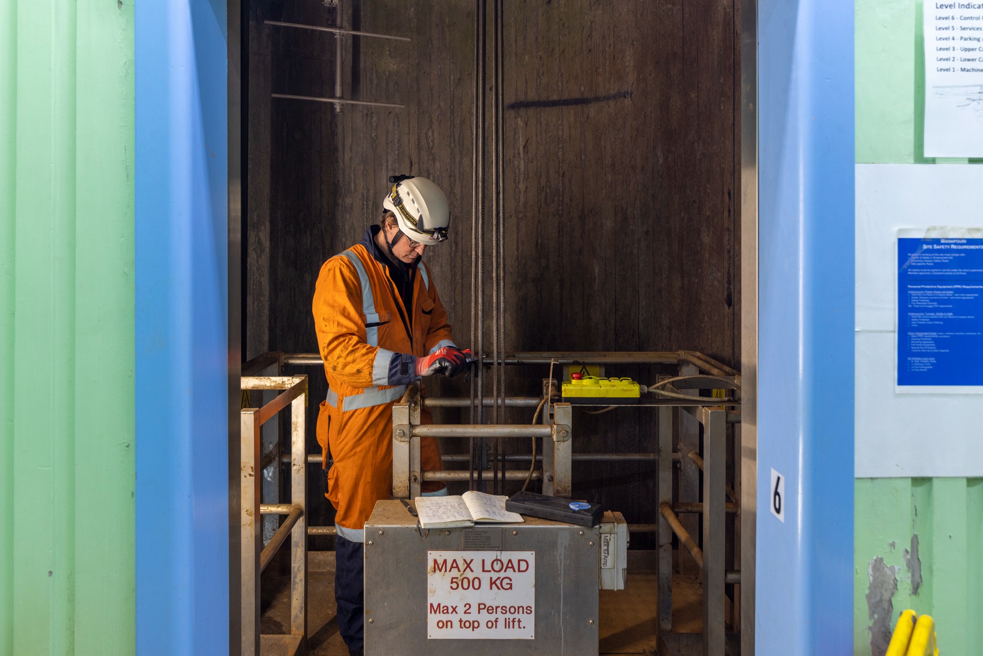 Pracownik w pomarańczowym kombinezonie i kasku z lampką, kontroluje liny w szybie dźwigu, z otwartym notatnikiem i żółtym panelem sterowania obok.