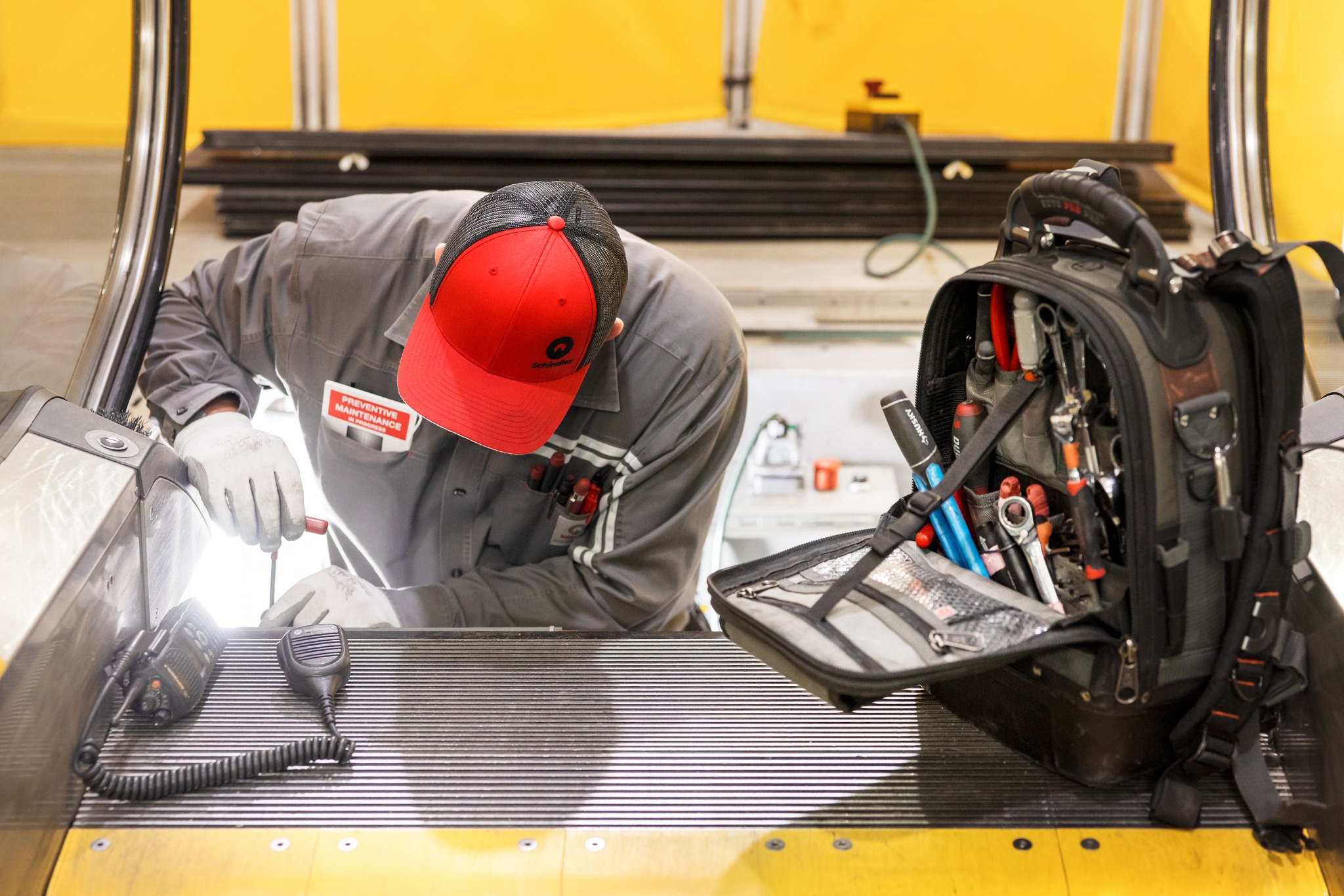 Technik w szarym kombinezonie i czerwonej czapce z daszkiem, pochylony nad naprawą windy, obok torba narzędziowa i radiotelefon.
