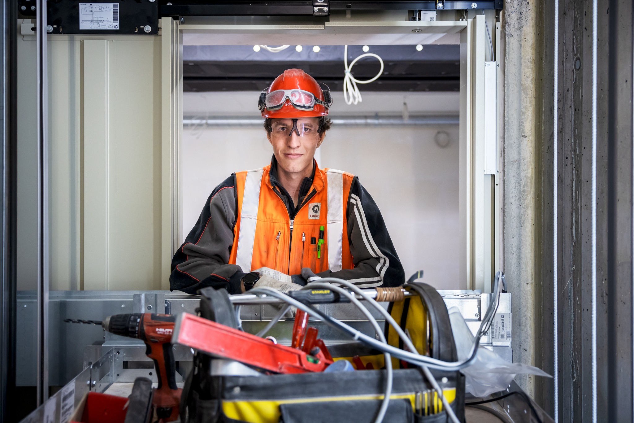 Pracownik w kasku i okularach ochronnych, w szybie windy w Warszawie, z narzędziami w skrzynce na pierwszym planie, podczas montażu lub serwisu.
