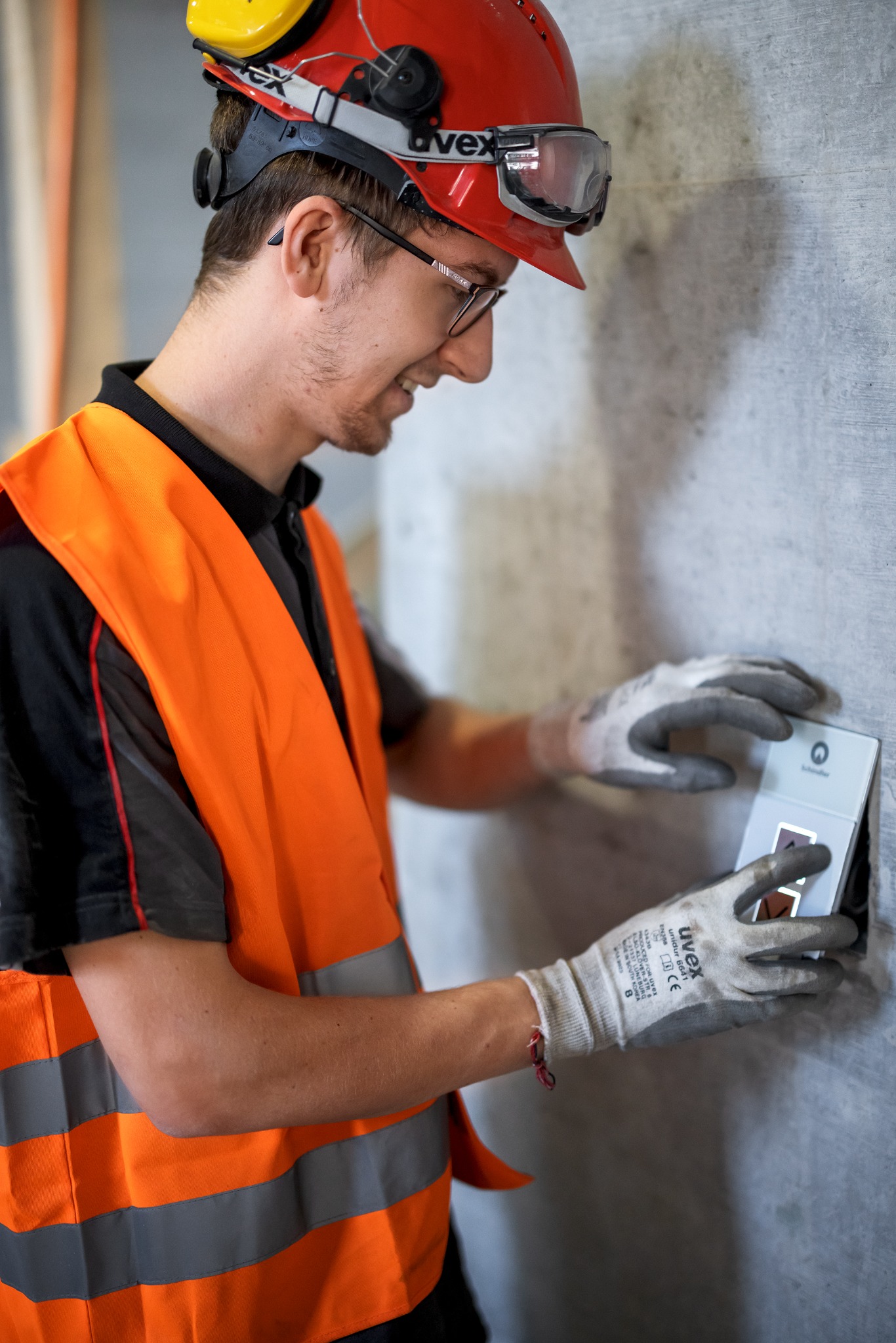 Mężczyzna w kasku i kamizelce odblaskowej, z uśmiechem, używa urządzenia pomiarowego marki Schindler na betonowej ścianie.
