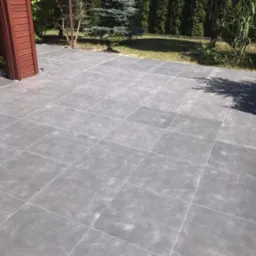 Szara, kwadratowa podłoga z płytek na zewnątrz, częściowo zacieniona, z zielonym ogrodem w tle i zabawkami dziecięcymi schowanymi pod drewnianym elementem konstrukcyjnym.