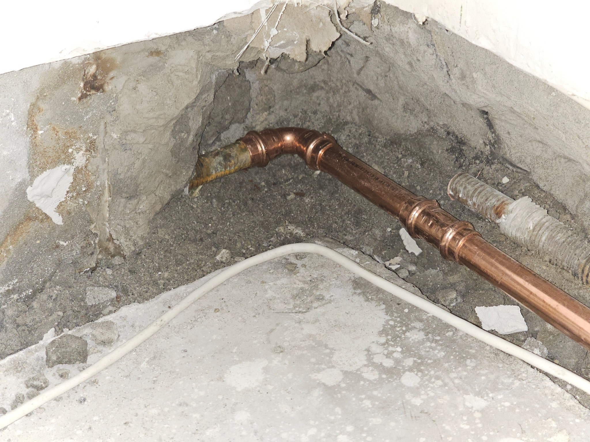 Miedziana instalacja centralnego ogrzewania z prasowanymi złączkami, częściowo ukryta w surowej ścianie z widocznymi śladami napraw, z fragmentem białego przewodu elektrycznego na pierwszym planie.